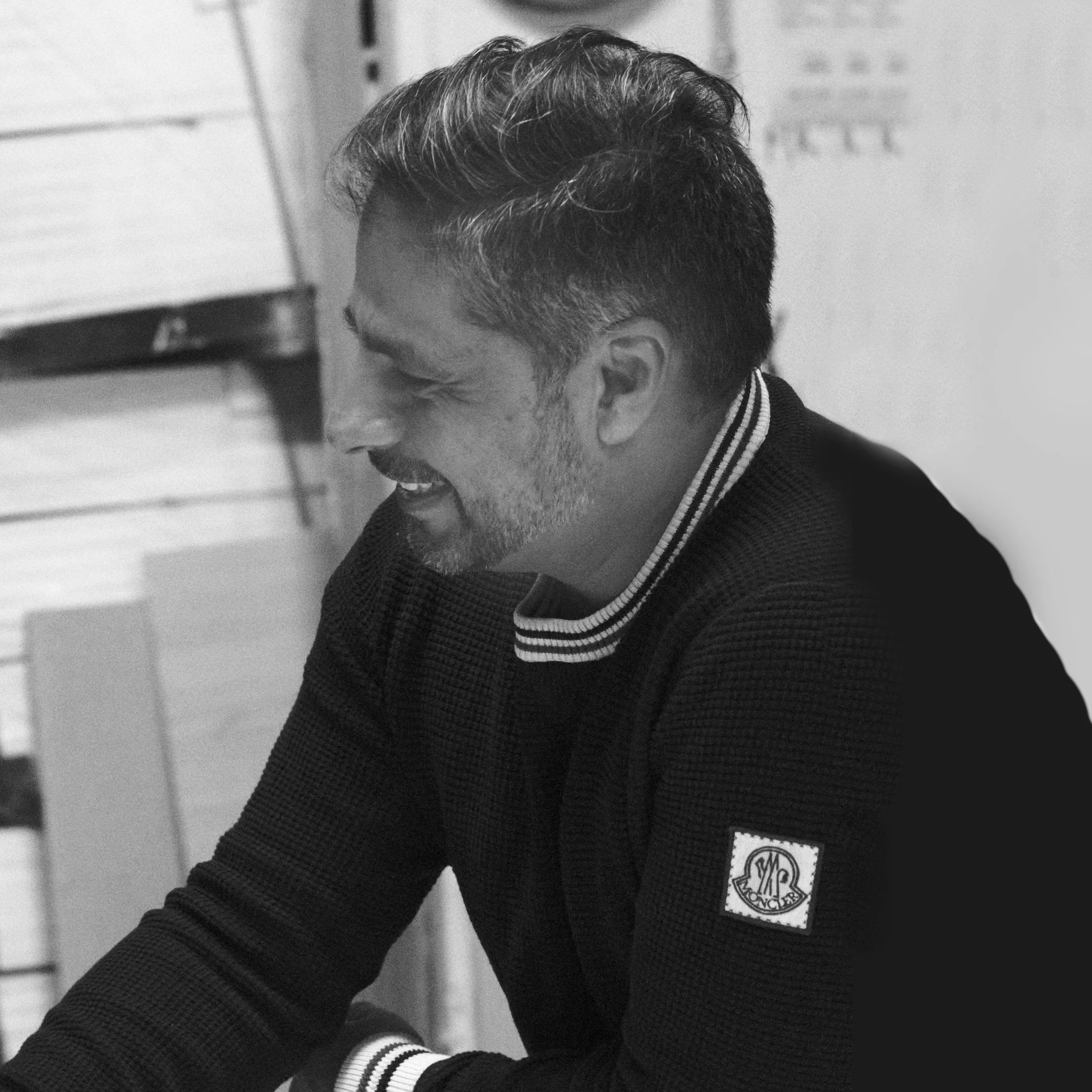 Un homme souriant avec des cheveux courts, portant un pull noir avec un logo Moncler sur la manche, photographié en noir et blanc.