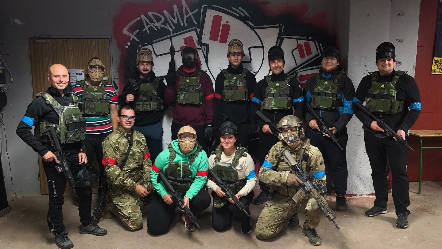 Group of 13 people in tactical gear posing with guns indoors at a paintball or airsoft facility, some wearing masks and camouflage, with a painted mural in the background.