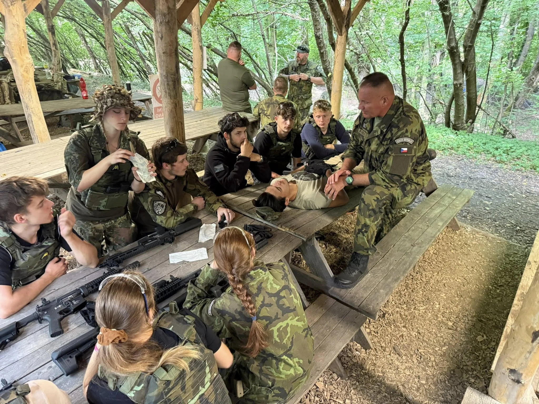 A group of young adults and military personnel gathered around a wooden picnic table in a wooded outdoor setting. Some are wearing camouflage uniforms, and one woman is dressed in a camouflage hat and jacket. There are rifles on the table, and a man 