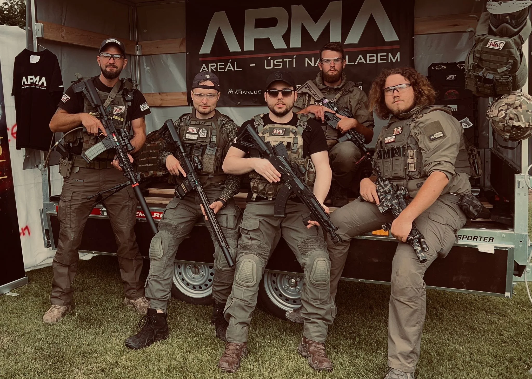 Six men wearing tactical gear and holding rifles pose in front of a booth with a banner that reads 'ARMA' and 'AREÁL - ÚSTÍ NAD LABEM.'