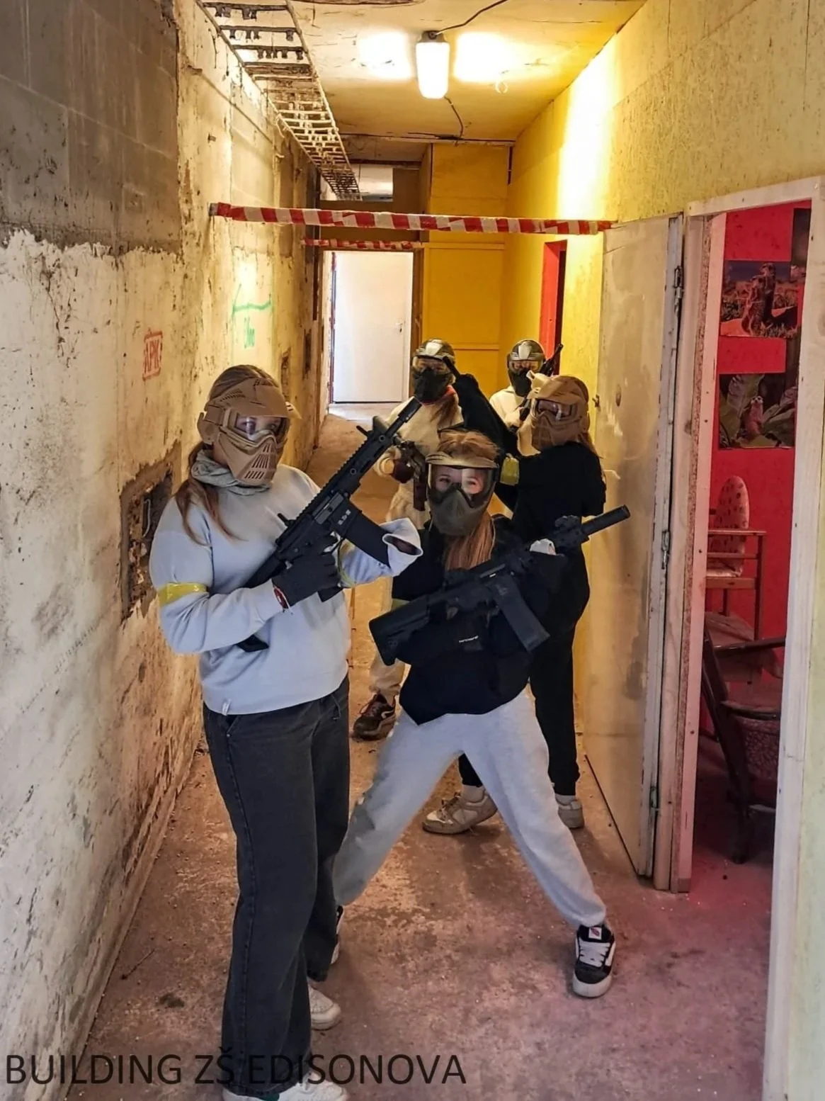 Group of people wearing paintball gear and holding guns inside an indoor paintball arena with yellow and beige walls.