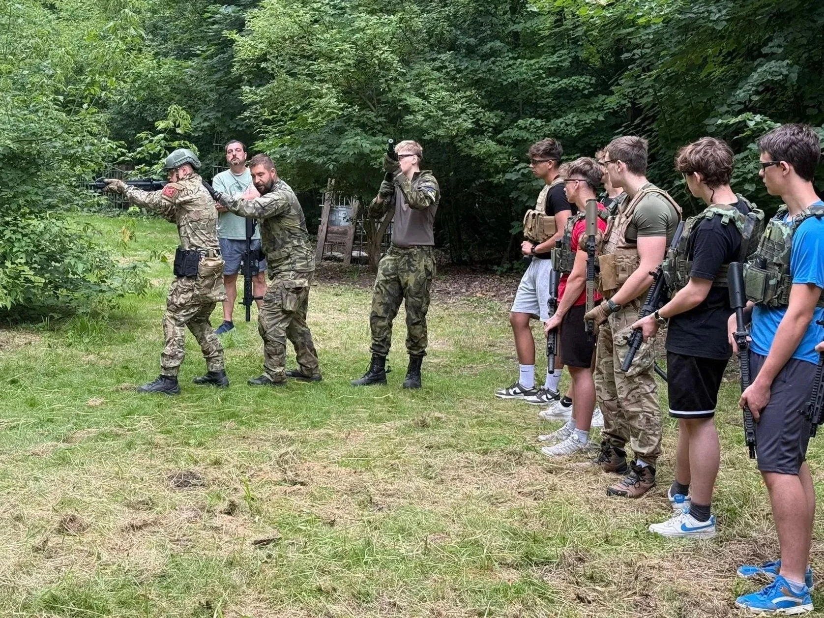 A group of young men, some in military gear, participating in a shooting or firearm training session outdoors in a wooded area, with two instructors guiding them.