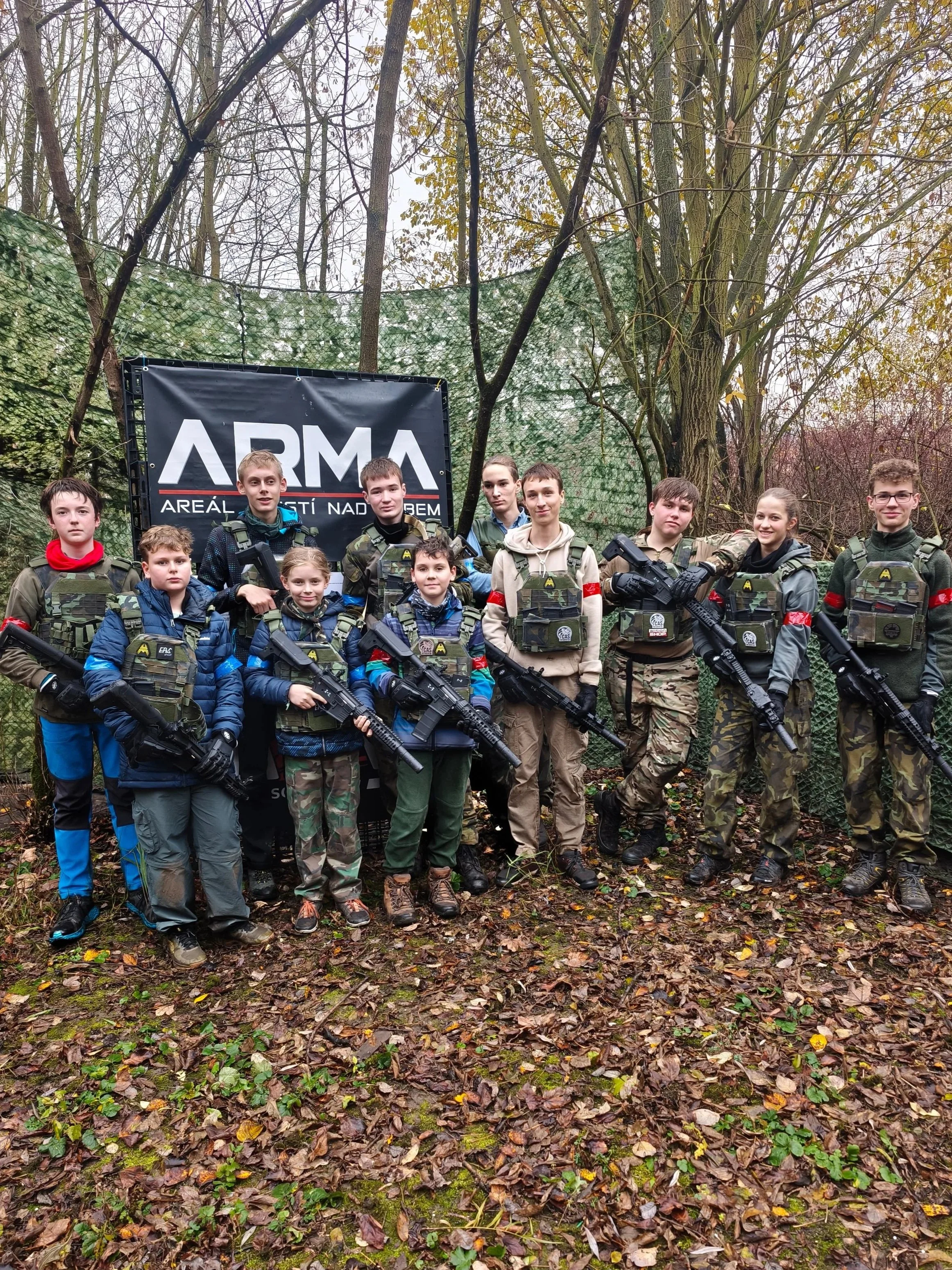 Group of children and teenagers in camouflage and tactical gear, holding toy guns, standing outdoors in wooded area with a large sign that reads 'ARMA' and outdoor netting behind them.