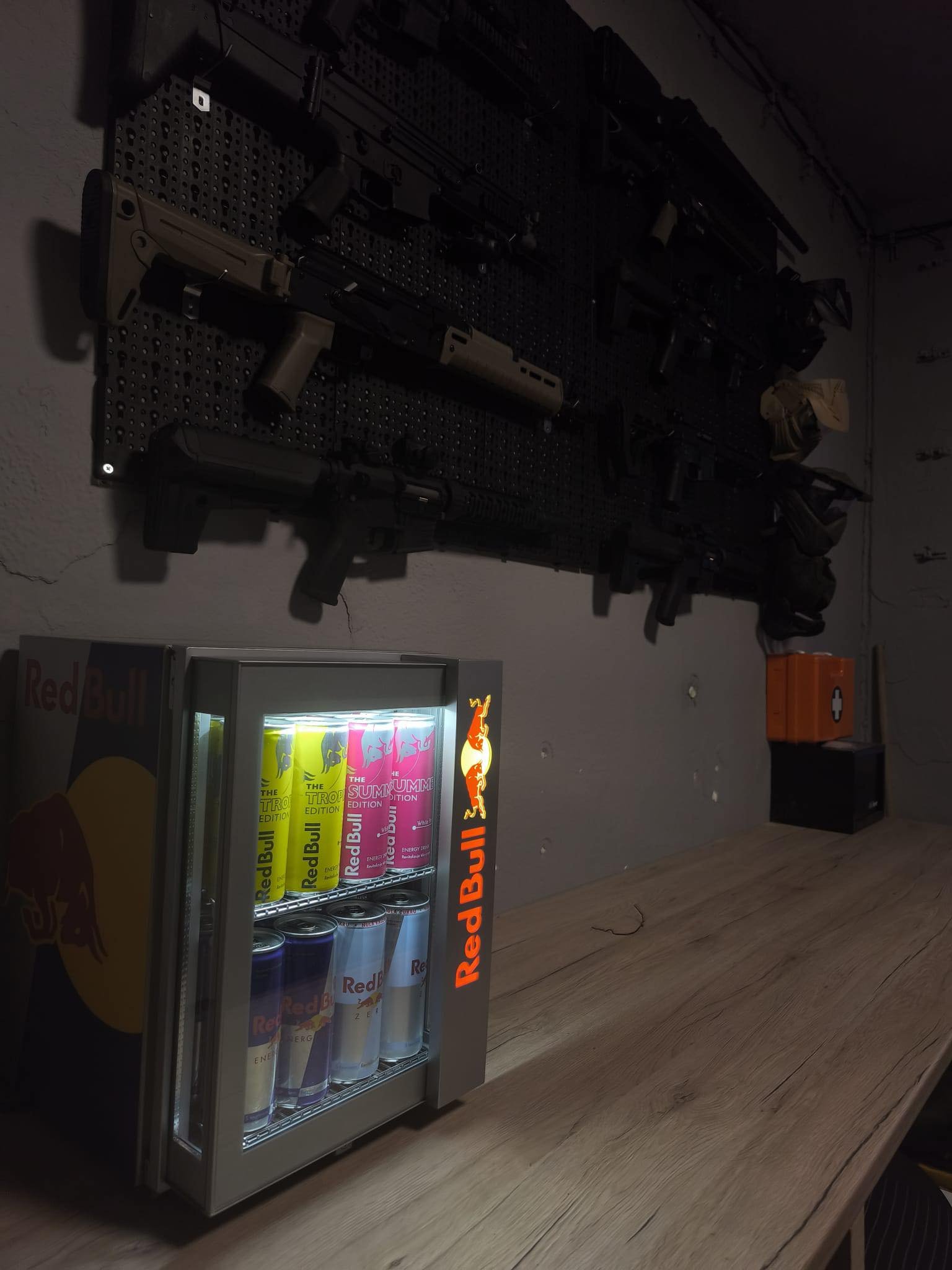 A mini refrigerator filled with Red Bull energy drinks sits on a wooden table in a dimly lit room. Guns are mounted on the wall above the table.