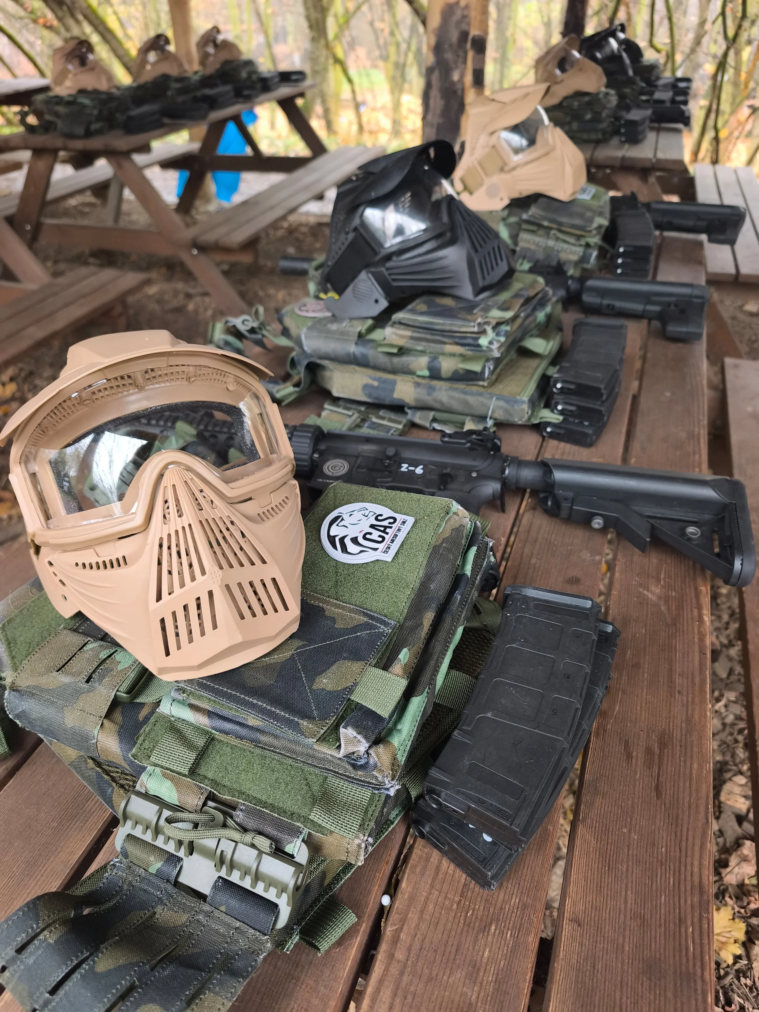 Paintball gear and equipment laid out on a wooden table, including masks, pistols, magazines, and camouflage gear in an outdoor setting.