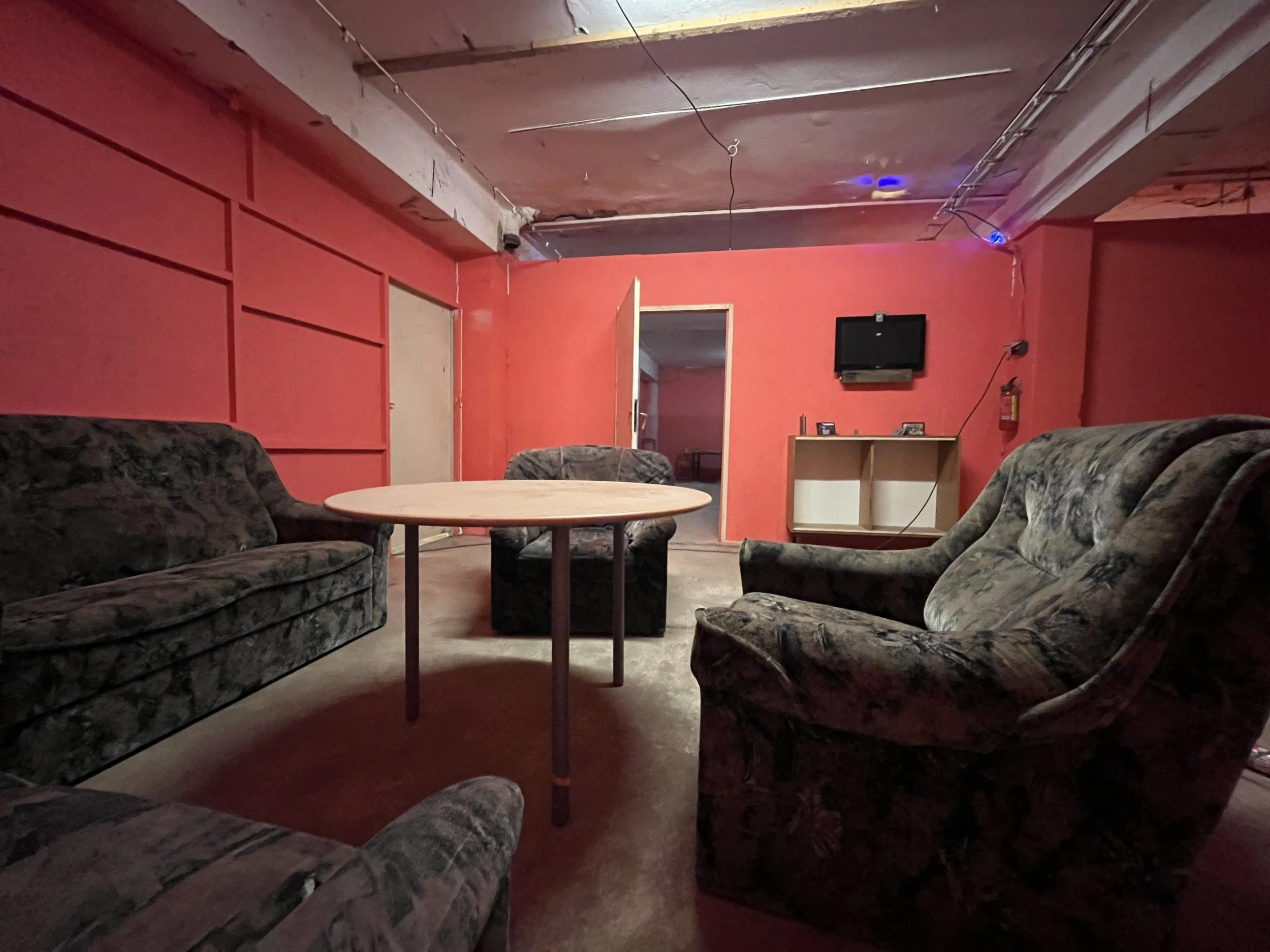 A dimly lit lounge room with red walls, a round wooden table, and three upholstered armchairs in dark patterned fabric. There is a small television mounted on the wall, a fire extinguisher, and a small shelf or cabinet underneath the TV. The ceiling shows exposed wiring and a blue light, with a partially open door leading to another room.