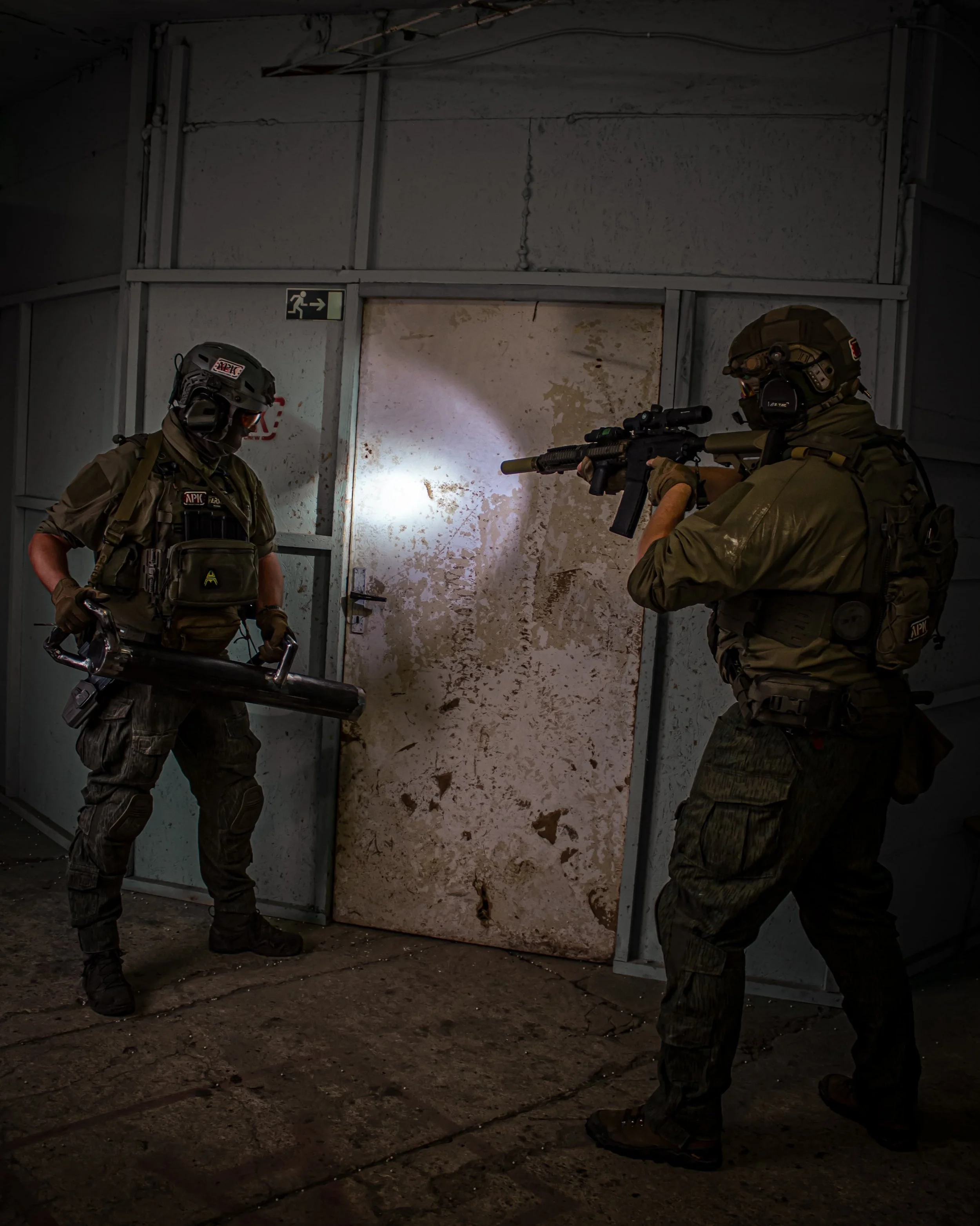 Two tactical armed officers in combat gear aiming at a door in a dimly lit indoor space.