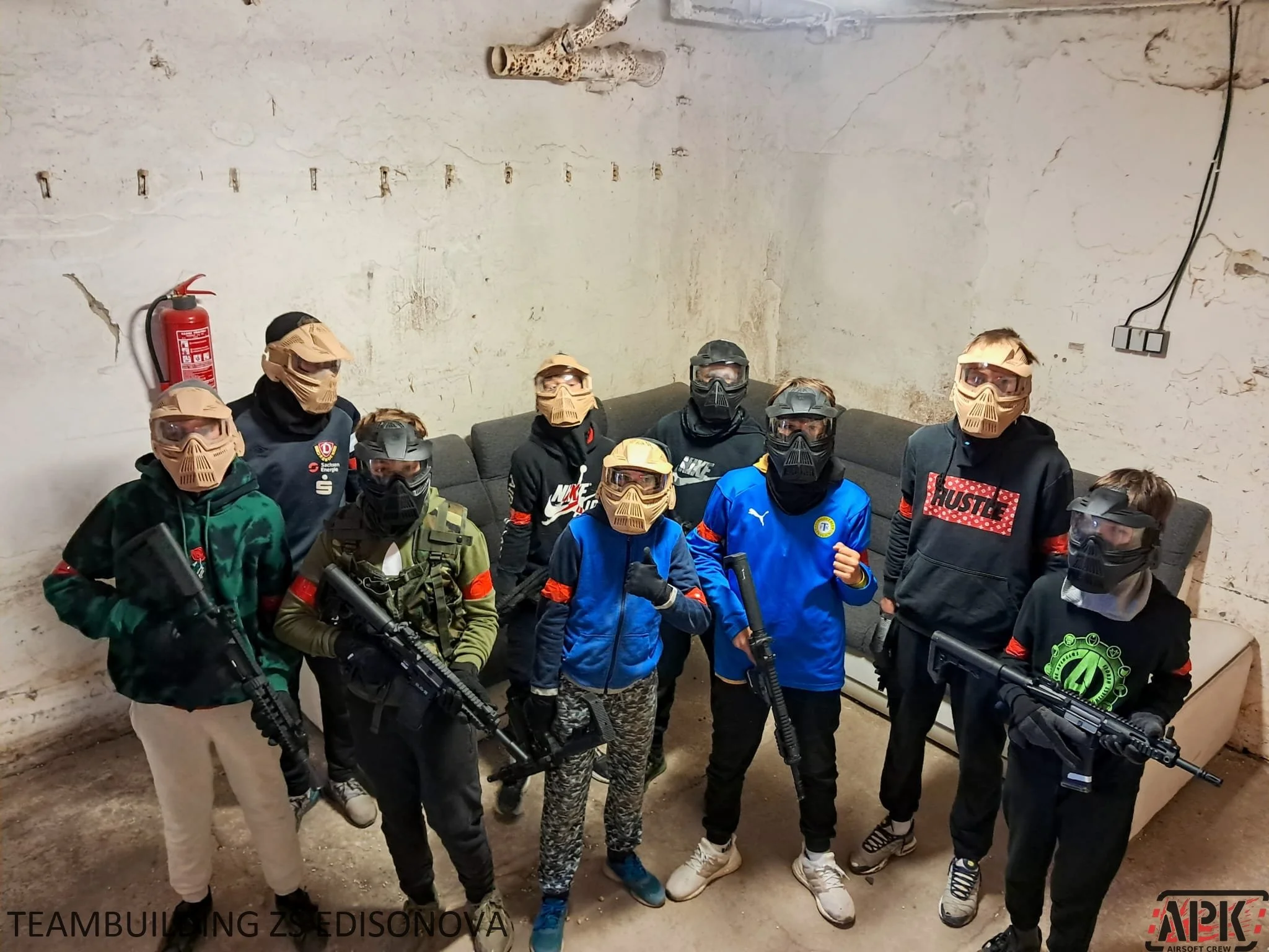 A group of people wearing tactical clothing, masks, and goggles, holding replica guns, in a room with a worn wall and a couch in the background.