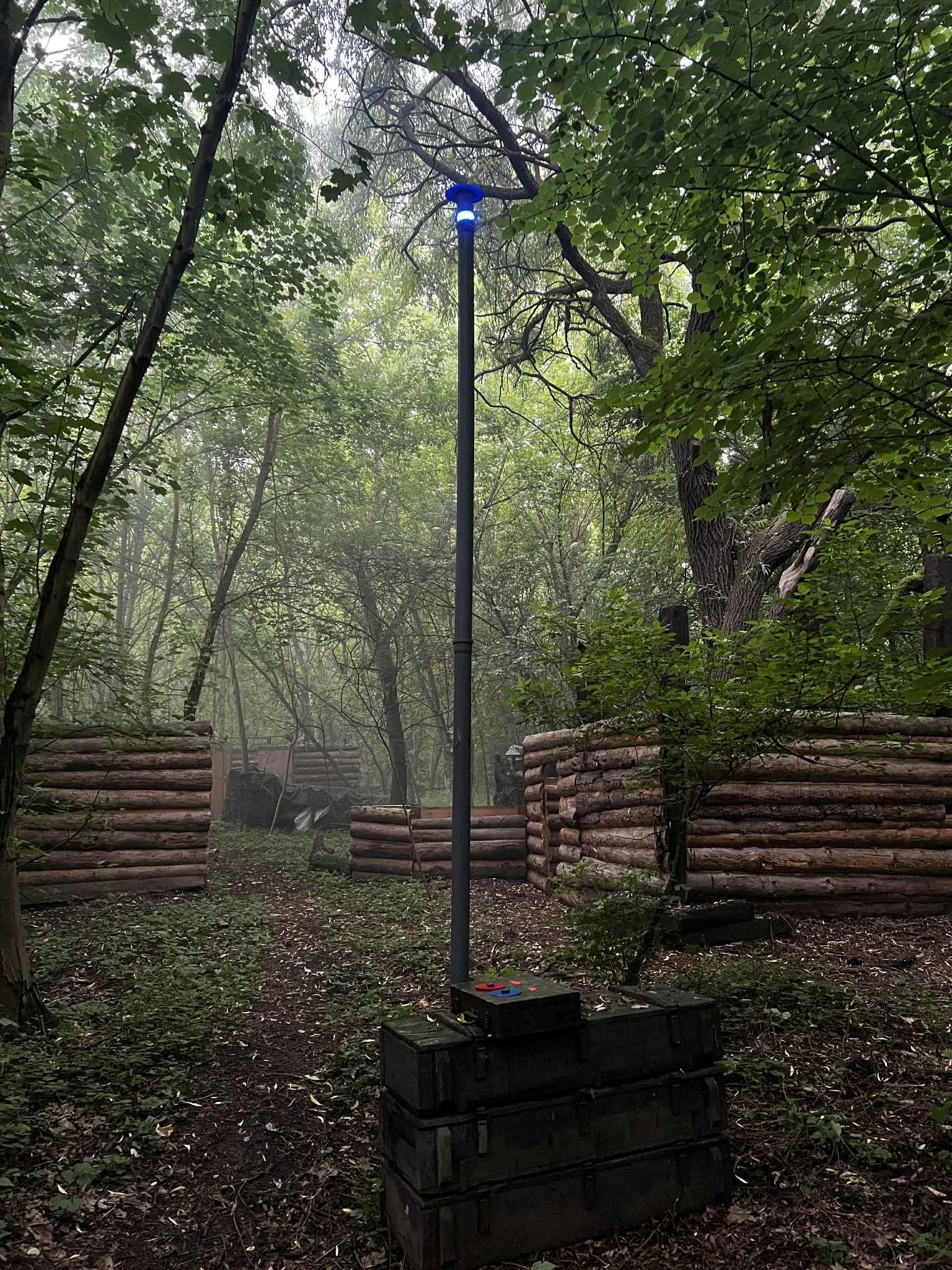 Tall pole with blue light on top standing on a black container in a wooded area with trees and log structures in the background.