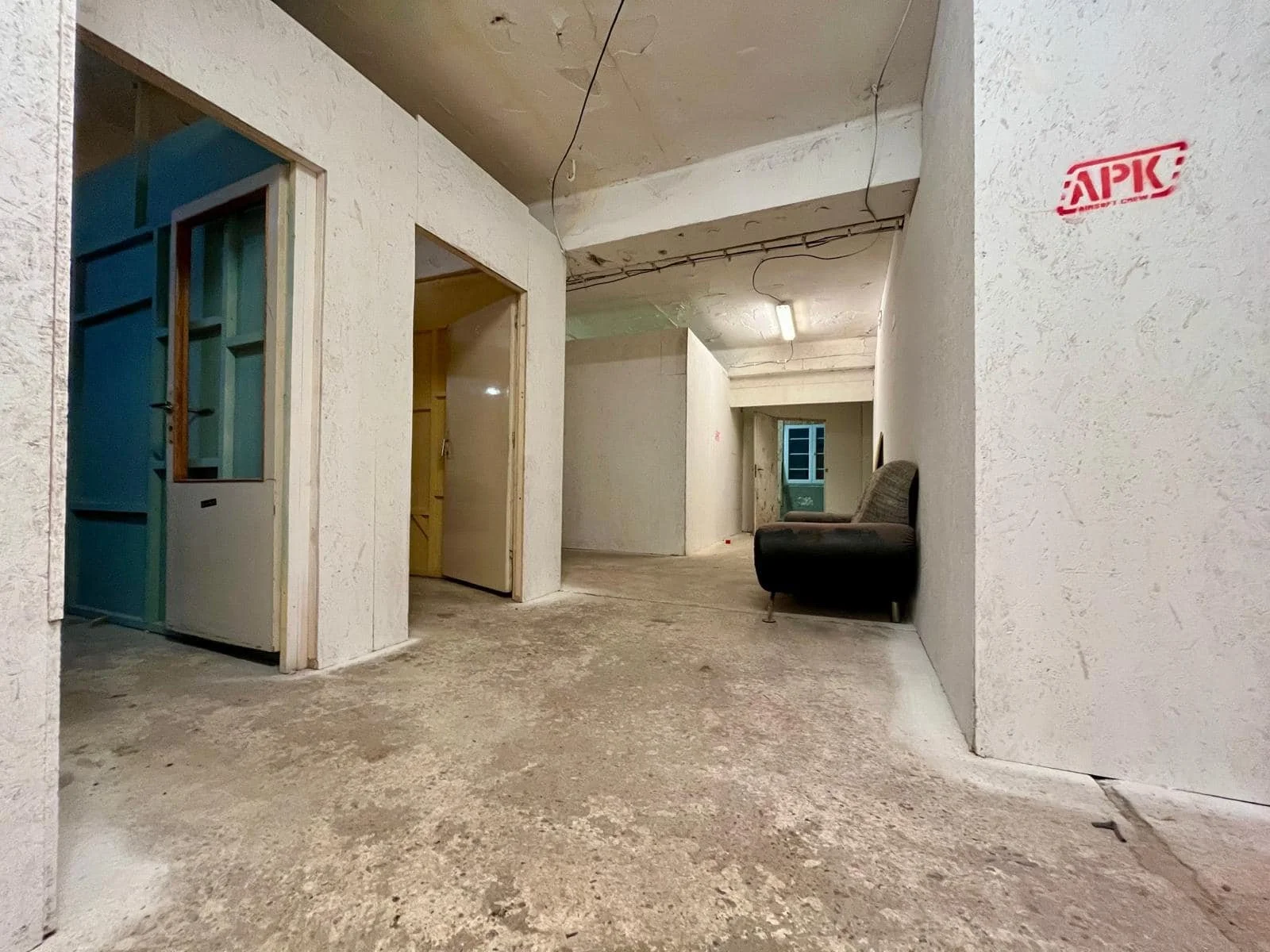 An unfinished interior space with exposed ceiling wiring, concrete floor, white textured walls, a black armchair against the right wall, and a doorway on the left side leading to another room.