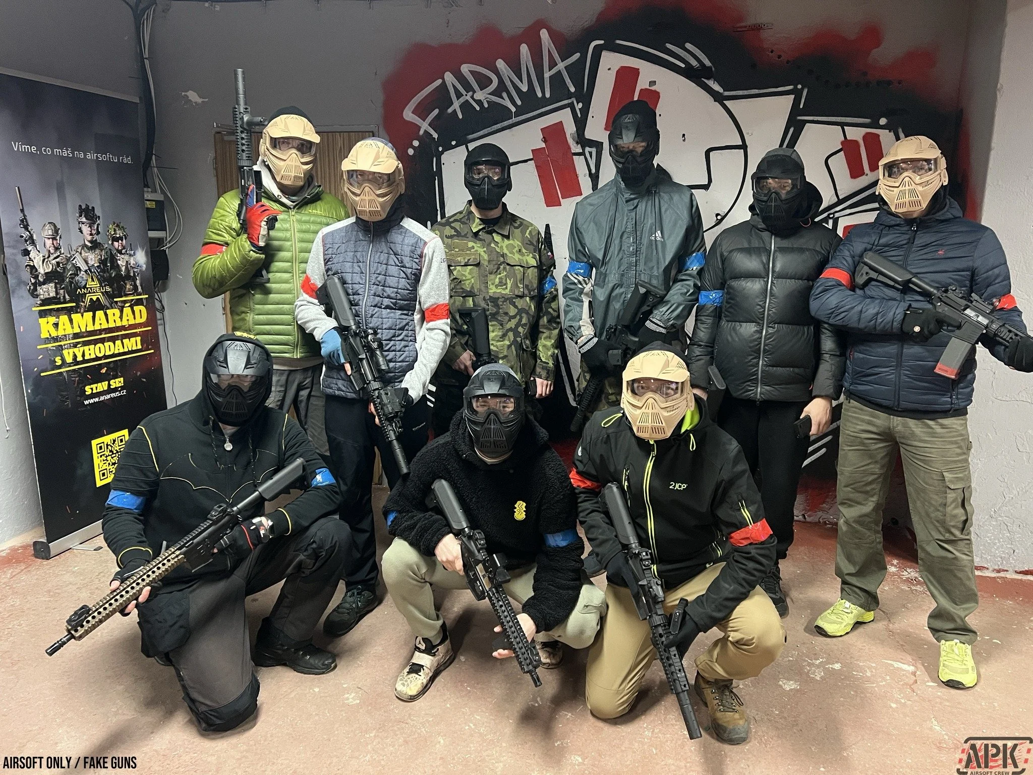 A group of ten people wearing tactical gear, masks, and helmets, posing indoors with airsoft guns. They are in front of a graffiti wall and a promotional poster on the left side.