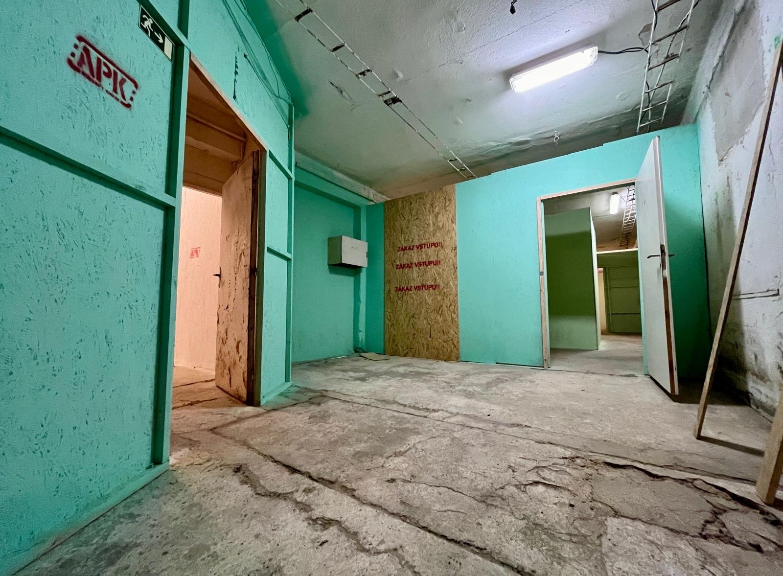 Interior view of an unfinished building with turquoise painted walls, open doorways leading to other rooms, a chair propped against wall, exposed wiring, and a concrete floor with cracks.