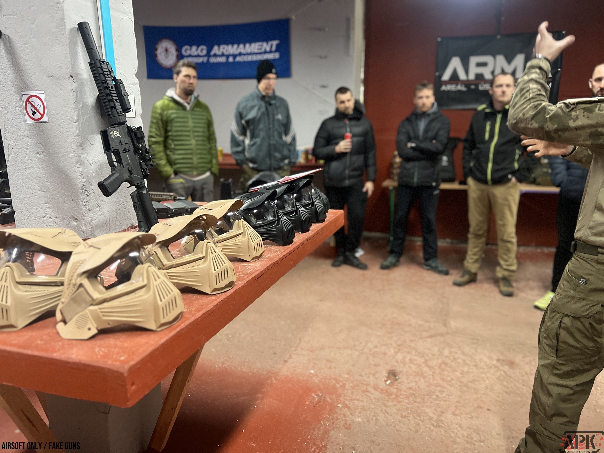 A table displaying a line of military-style face masks in tan and black, with a rifle leaning against a wall next to the table. In the background, a group of men is listening to a person giving a presentation. The setting appears to be an indoor fire