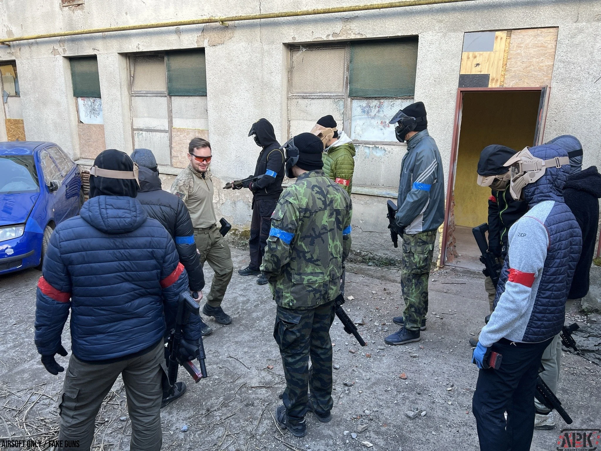 Group of people wearing tactical gear and masks standing outside near a weathered building with boarded-up windows.