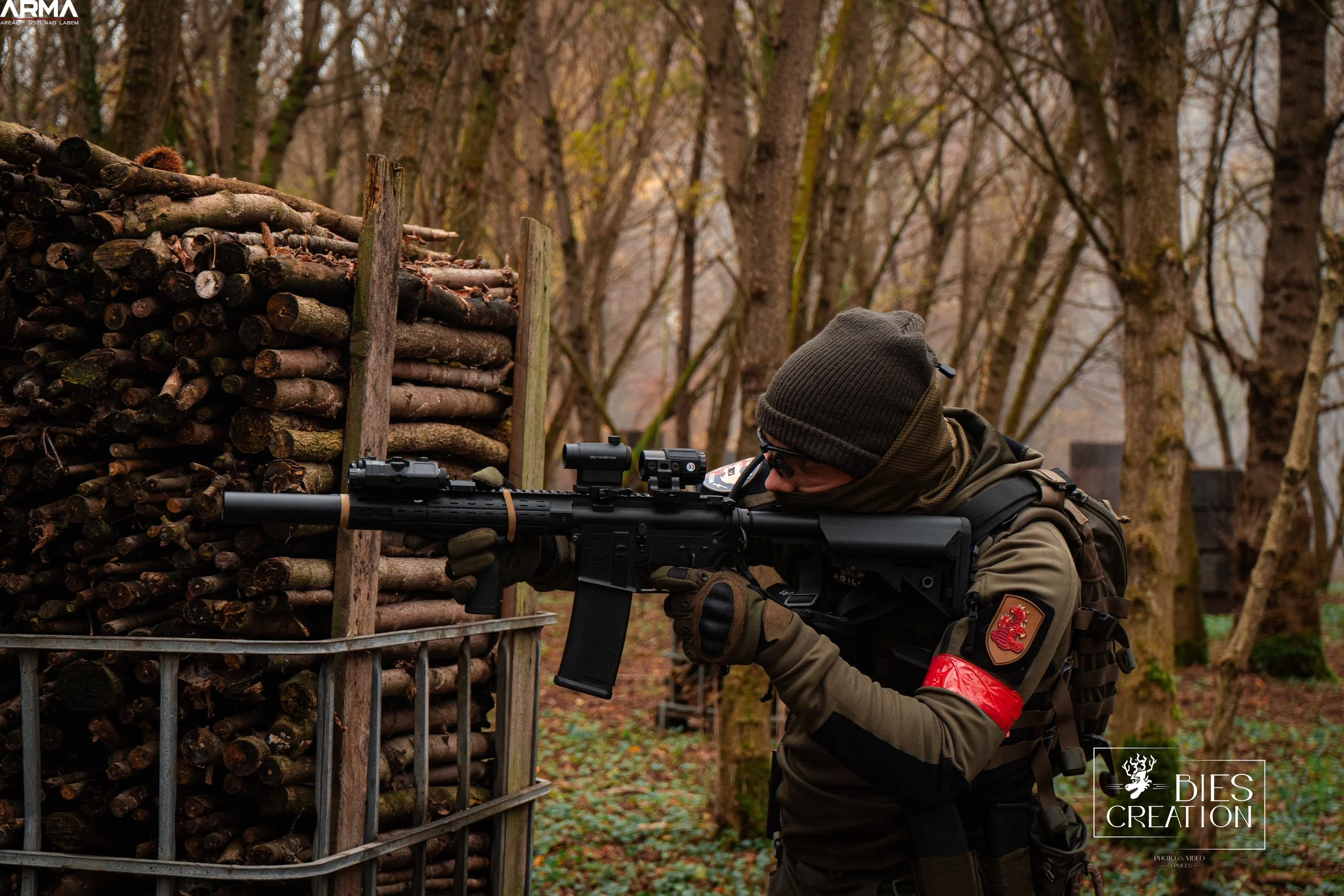 A person dressed in tactical gear aiming a rifle with a scope in a wooded area, crouched behind a wooden structure with stacked logs.