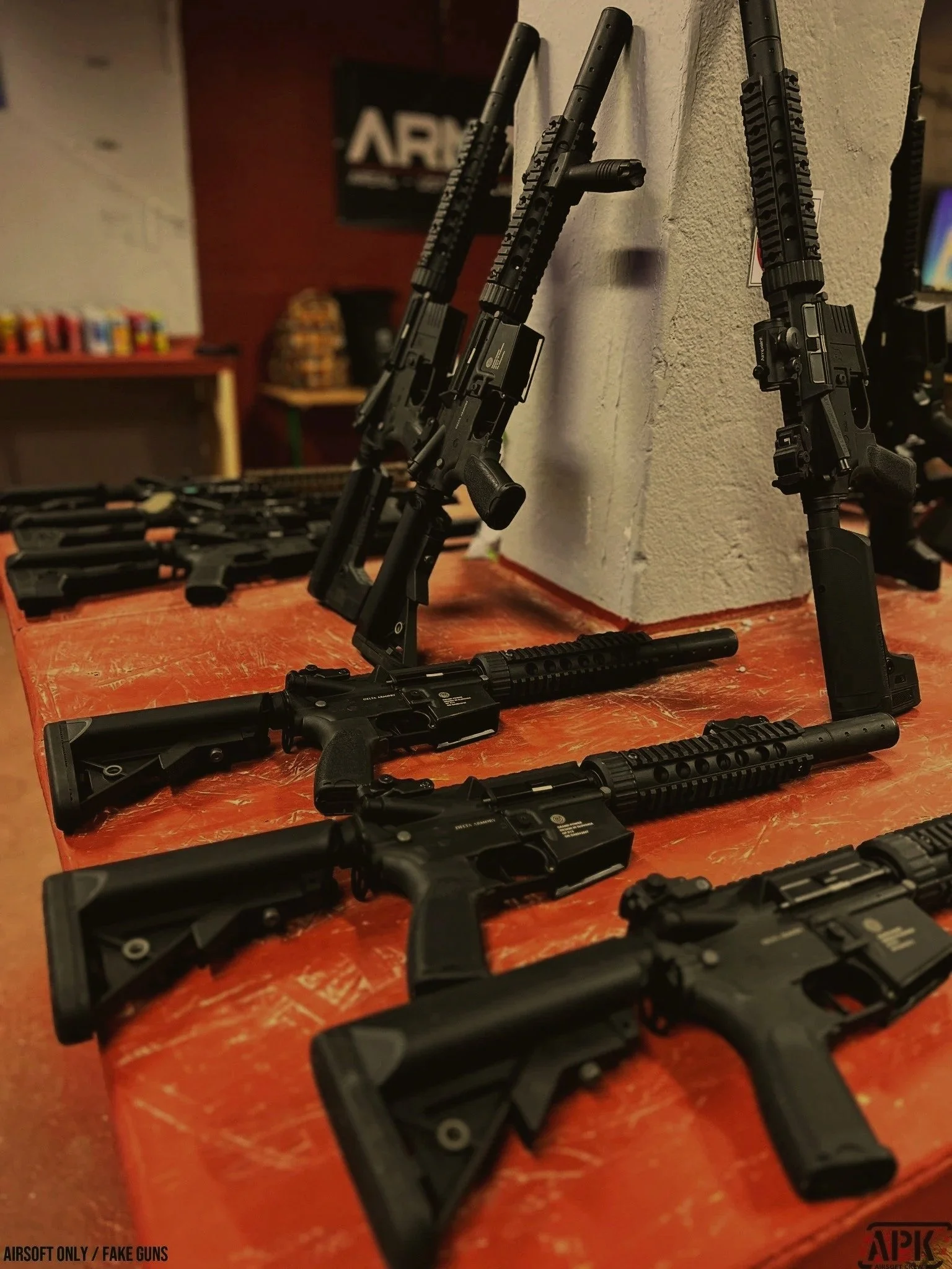 Multiple black rifles and handguns arranged on a table with a white wall and a poster in the background.