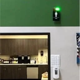 Hospital or medical facility corridor featuring a green exit sign, a black door monitoring system, and a hand sanitizer dispenser on the wall.
