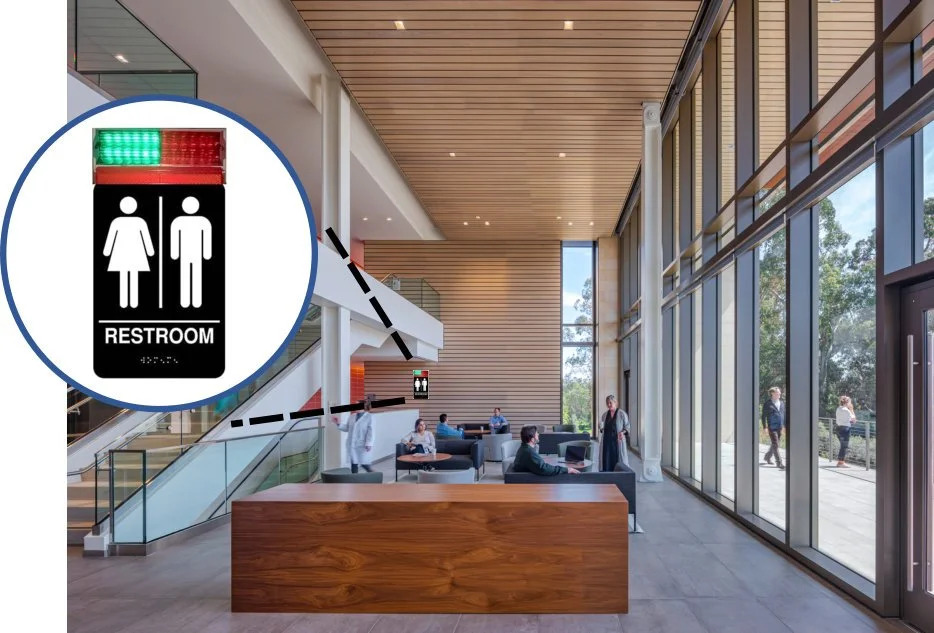 Lobby of a modern building with large glass windows, seating area, and a sign indicating the location of restrooms with male, female, and handicap symbols, and a green light at the top.