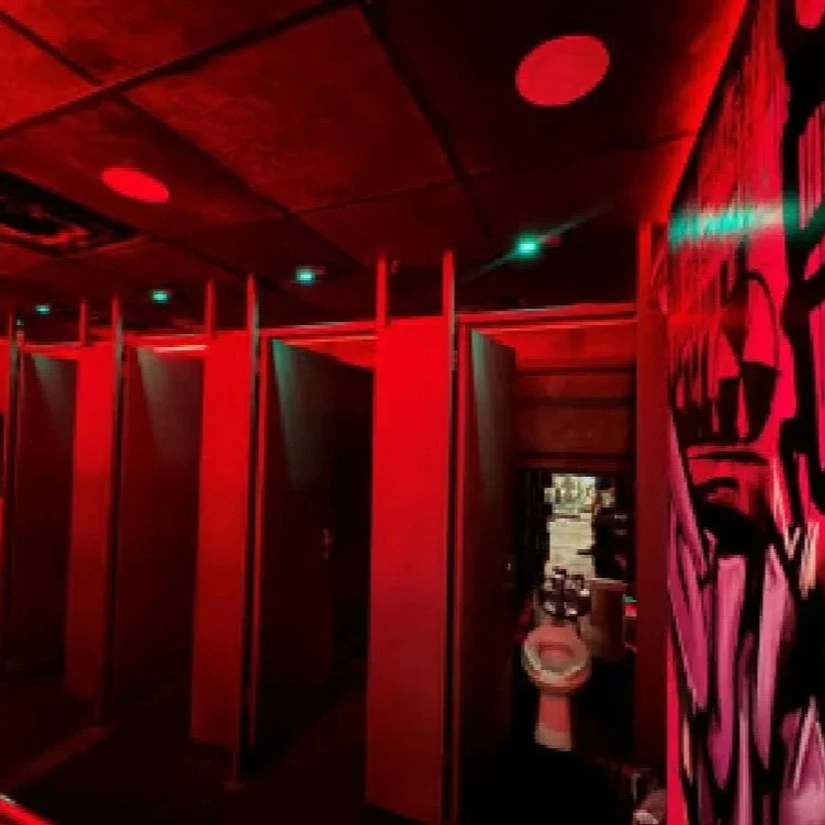 Public restroom with individual stalls illuminated by red lighting, tiled walls, and a graffiti mural on the right wall.
