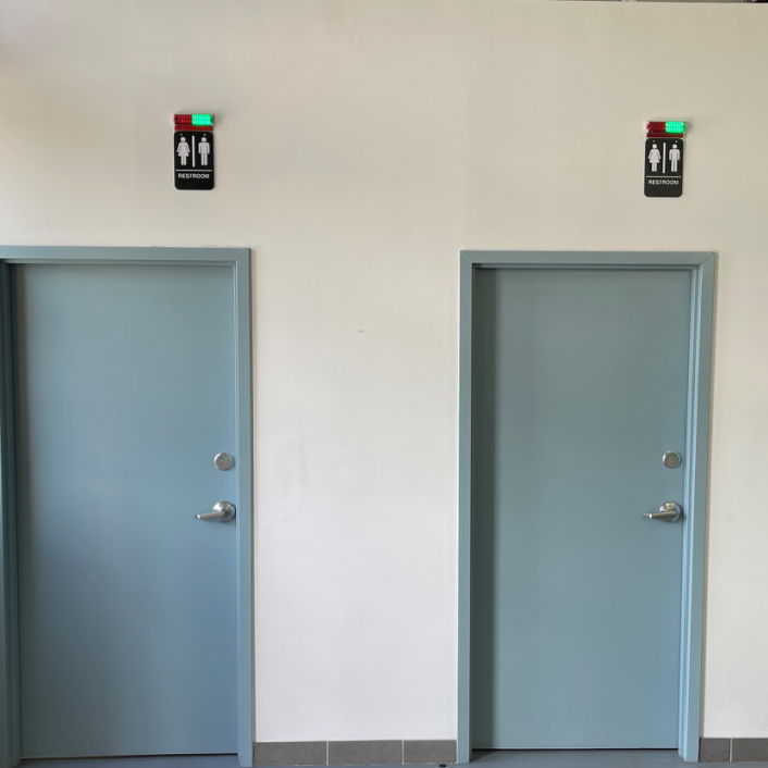 Two gray restroom doors with signs indicating men's and women's restrooms, mounted above each door, with one sign showing red and green lights.