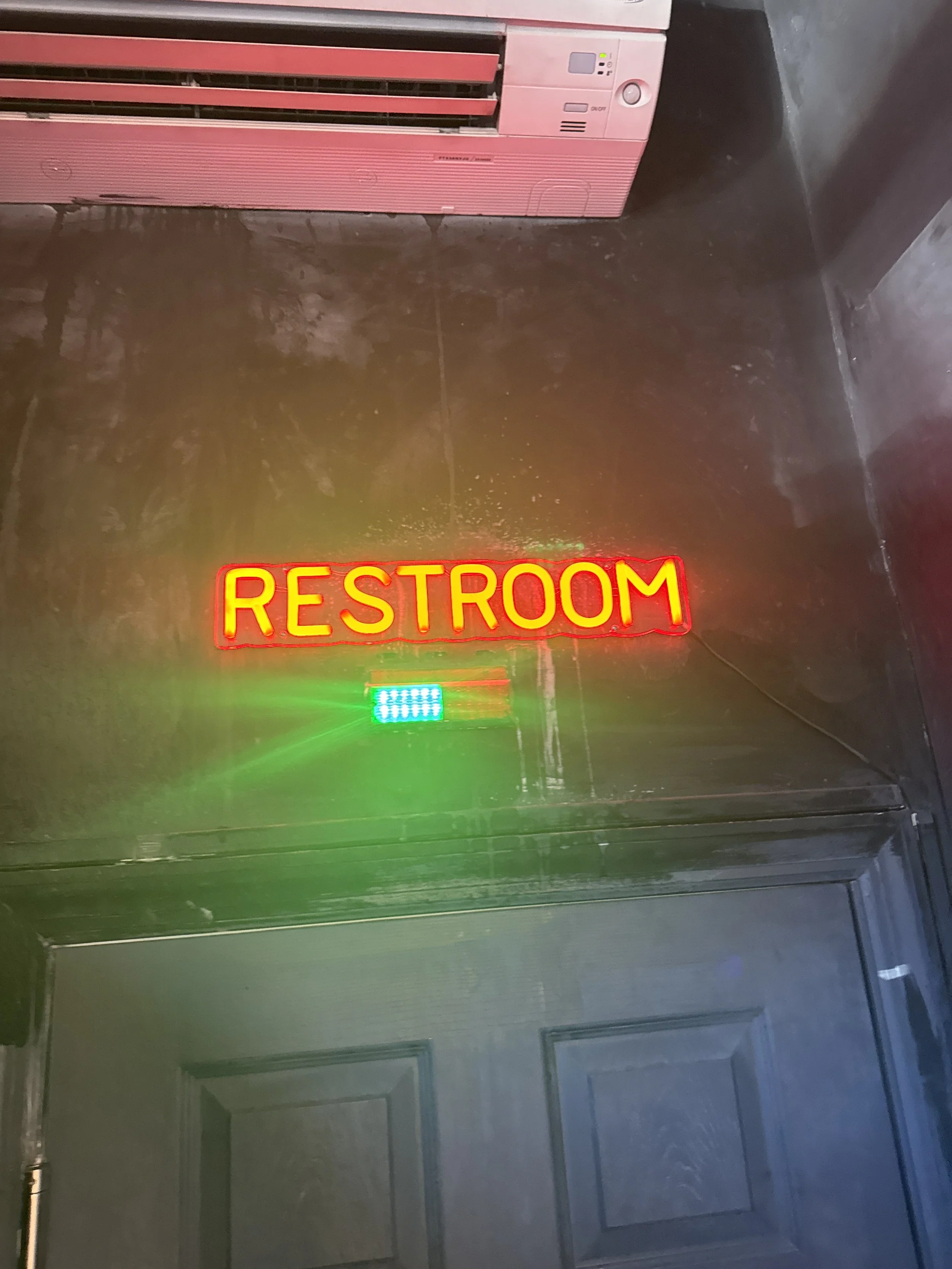 Pink air conditioning unit mounted on a wall above a dark-colored door with a neon red and yellow "RESTROOM" sign illuminated below it.
