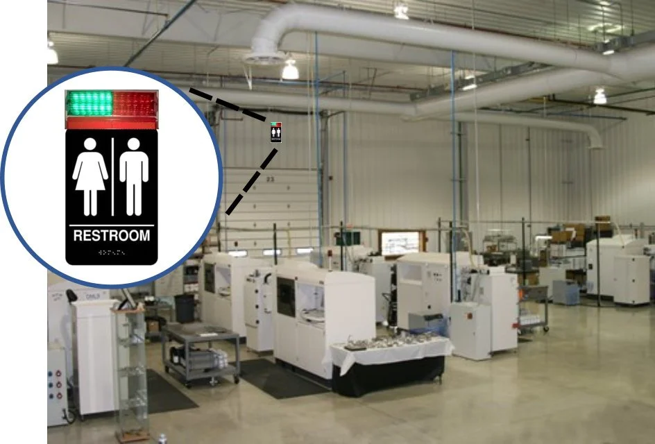 A sign indicating a restroom for men and women with a red and green light above it, hanging in an industrial or manufacturing space with machines and equipment in the background.