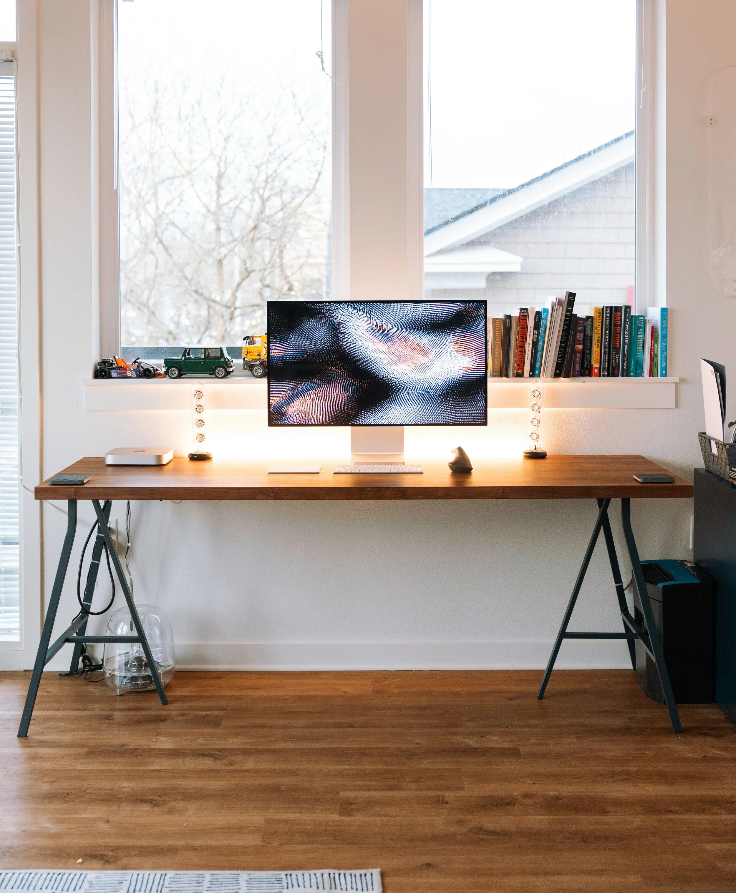 Home office desk with a computer monitor, books on the windowsill, toy cars, and a small lamp, set in front of a window with trees and a house roof visible outside.