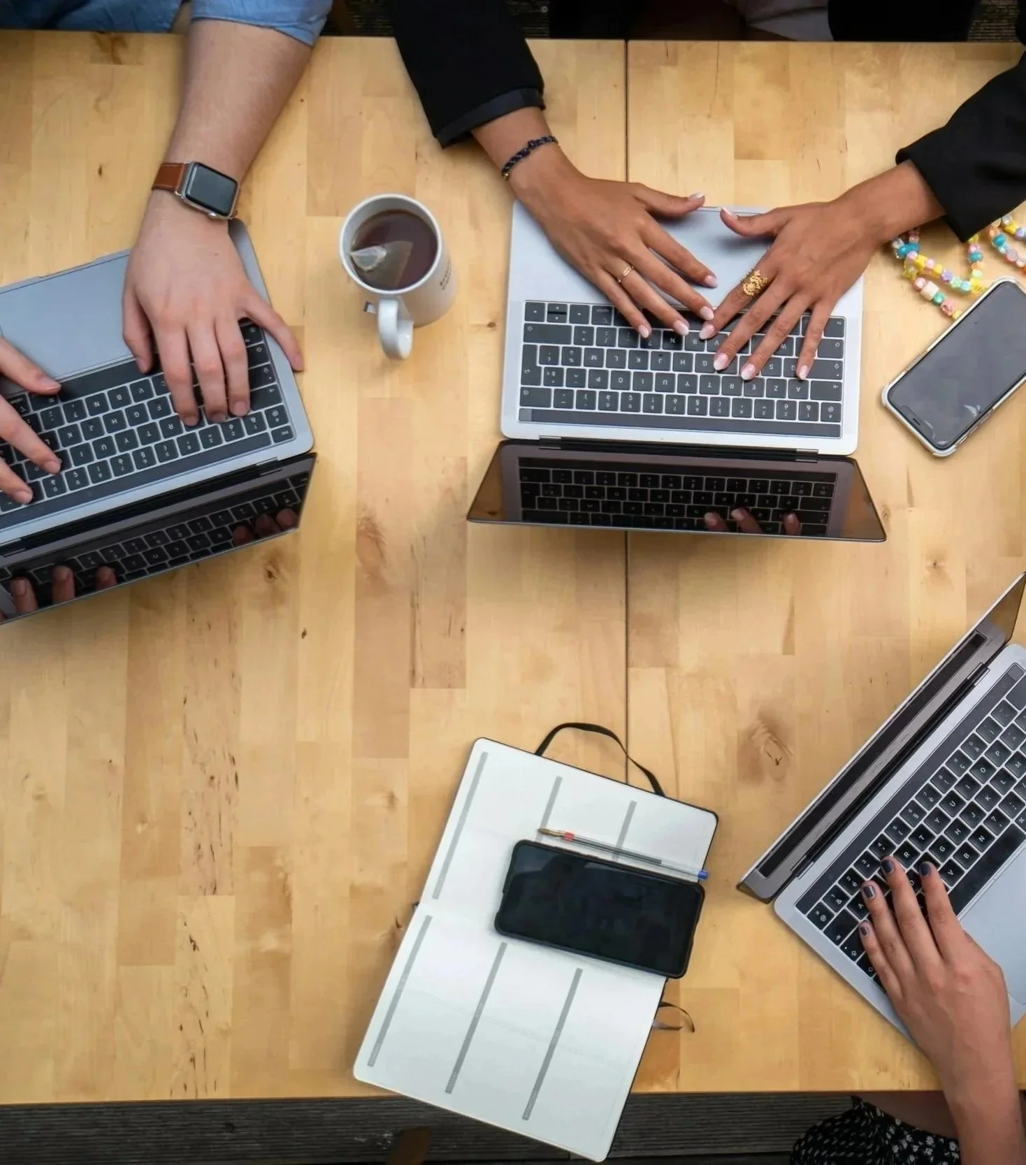 Top-down view of a wooden table with four people working on laptops, a mug of tea, a smartphone, a notepad with a phone and a few pens, and some beaded jewelry.