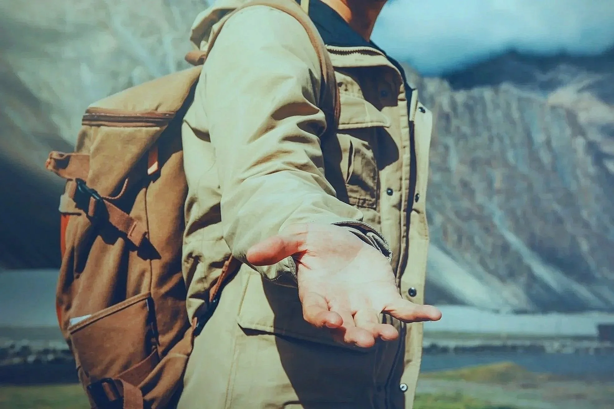 Person wearing outdoor gear and carrying a backpack, extending their hand, with mountainous landscape in the background.