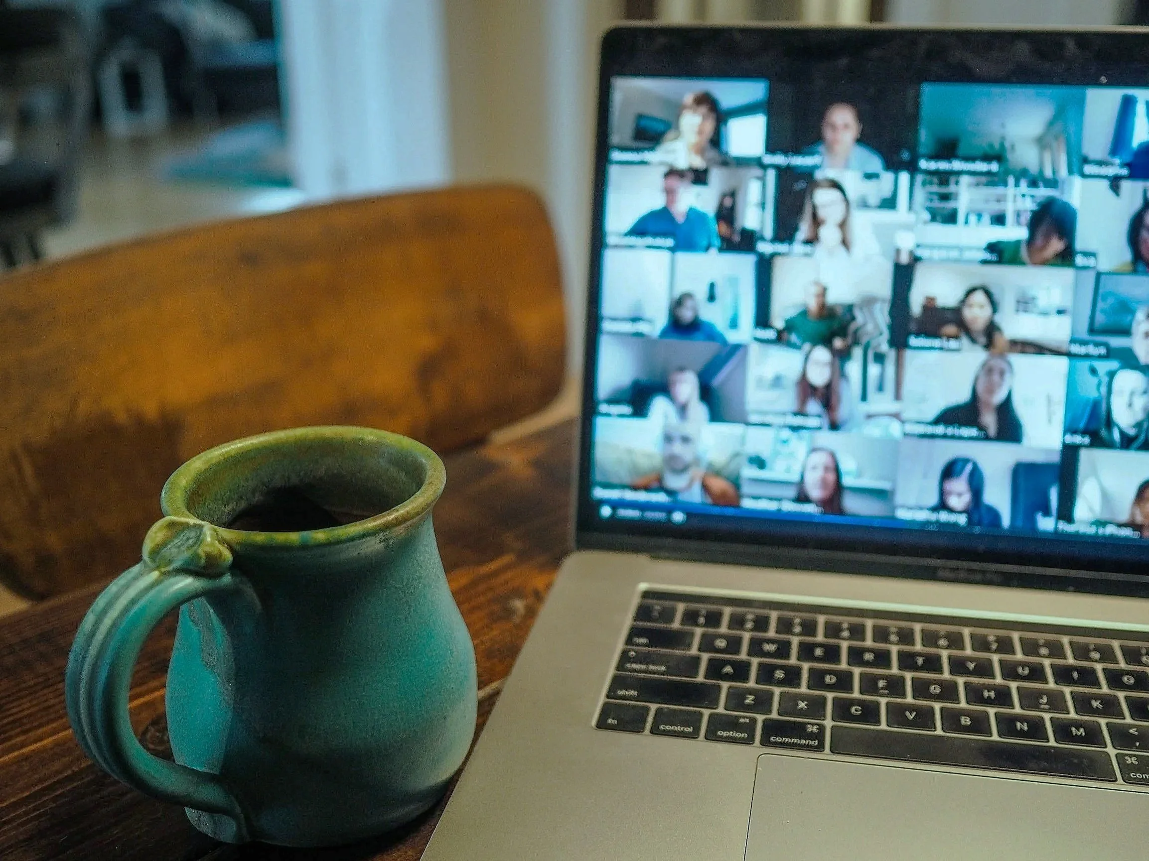 Person participating in a virtual meeting on a laptop screen with four colleagues, holding a coffee mug and taking notes in a bright office or home workspace.