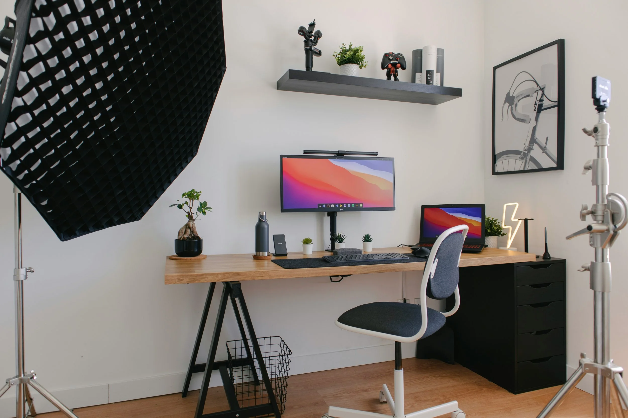 Home office setup with a wooden desk, dual monitors, potted plants, a laptop, desk lamp, and photo frame, with studio lighting equipment in a bright room.