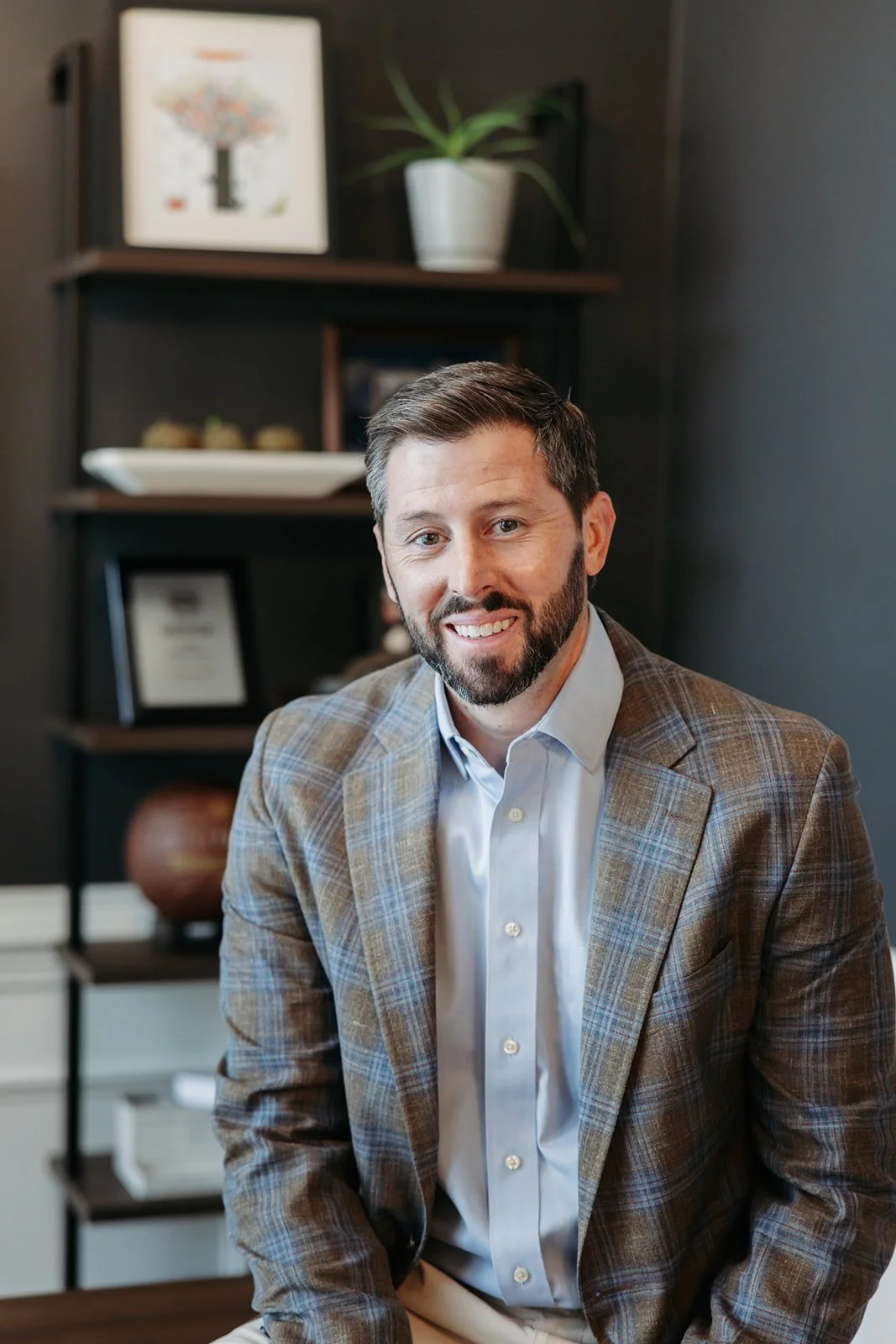 A man with a beard and short hair, dressed in a plaid blazer and light blue button-up shirt, sitting and smiling in an office or home office setting with dark walls and shelves with decor and framed items in the background.