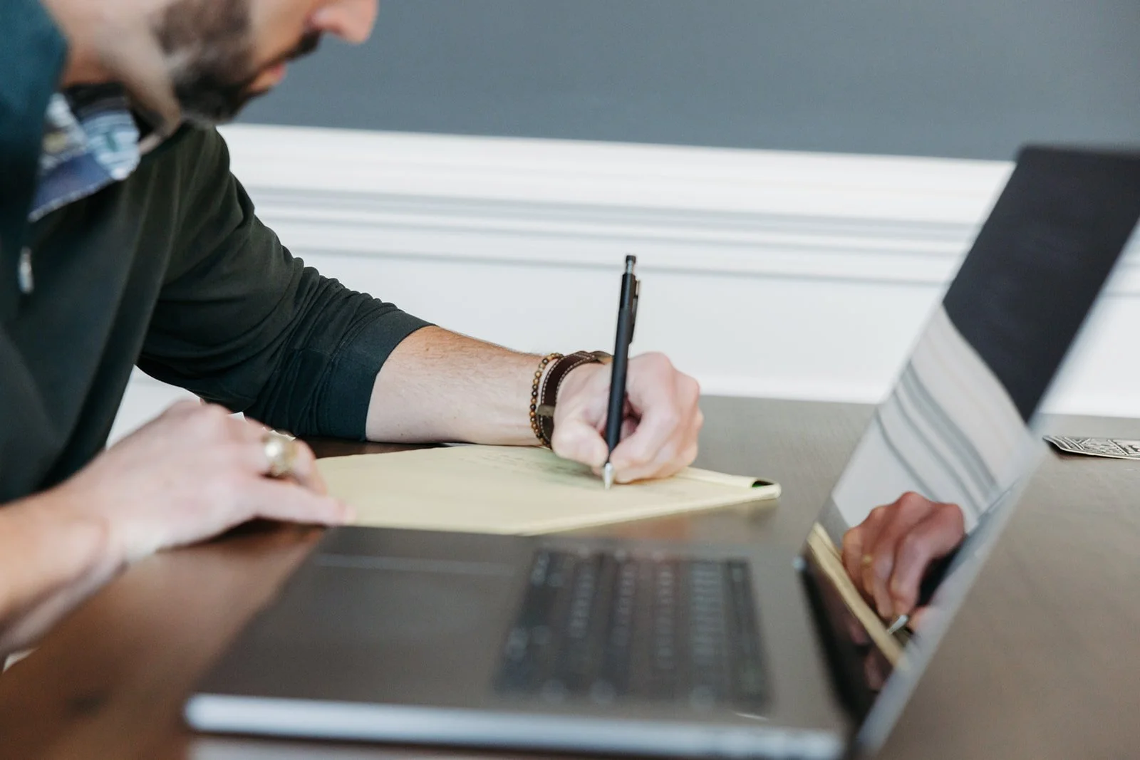 Person sitting at a desk writing on a yellow notepad with a pen, with a laptop open in front of them.