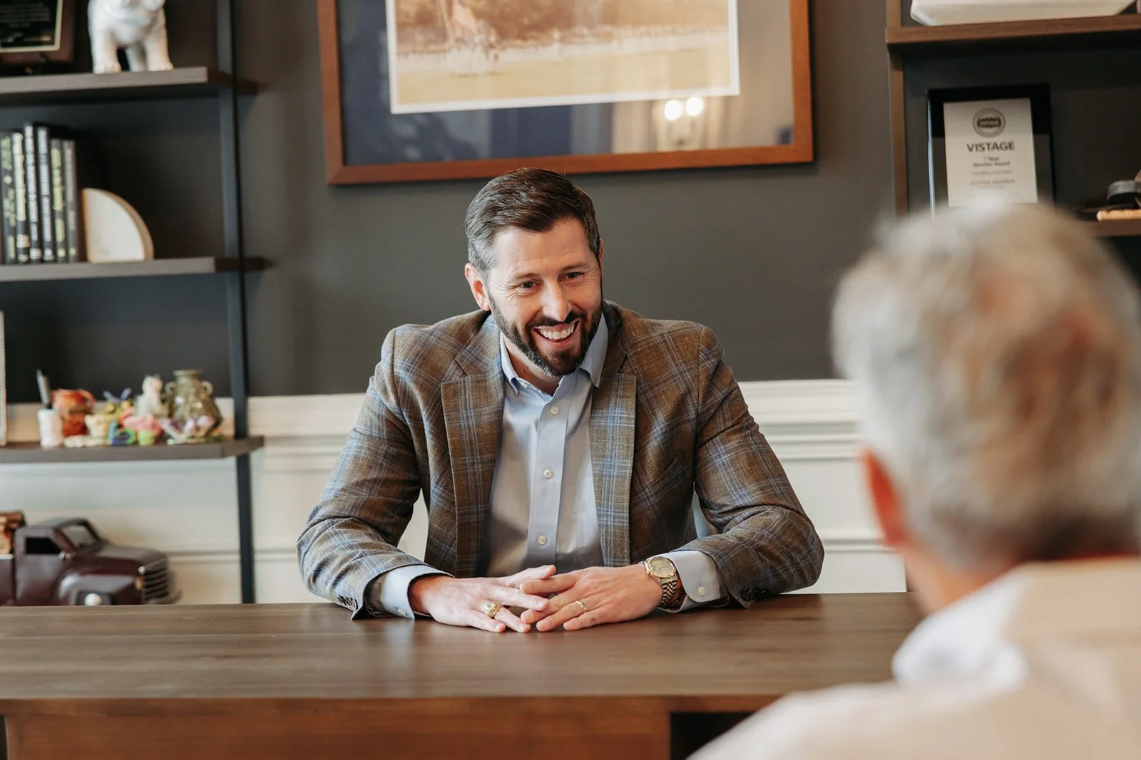 A man in a plaid suit smiling and talking to an elderly man at a wooden table in an office or conference room.