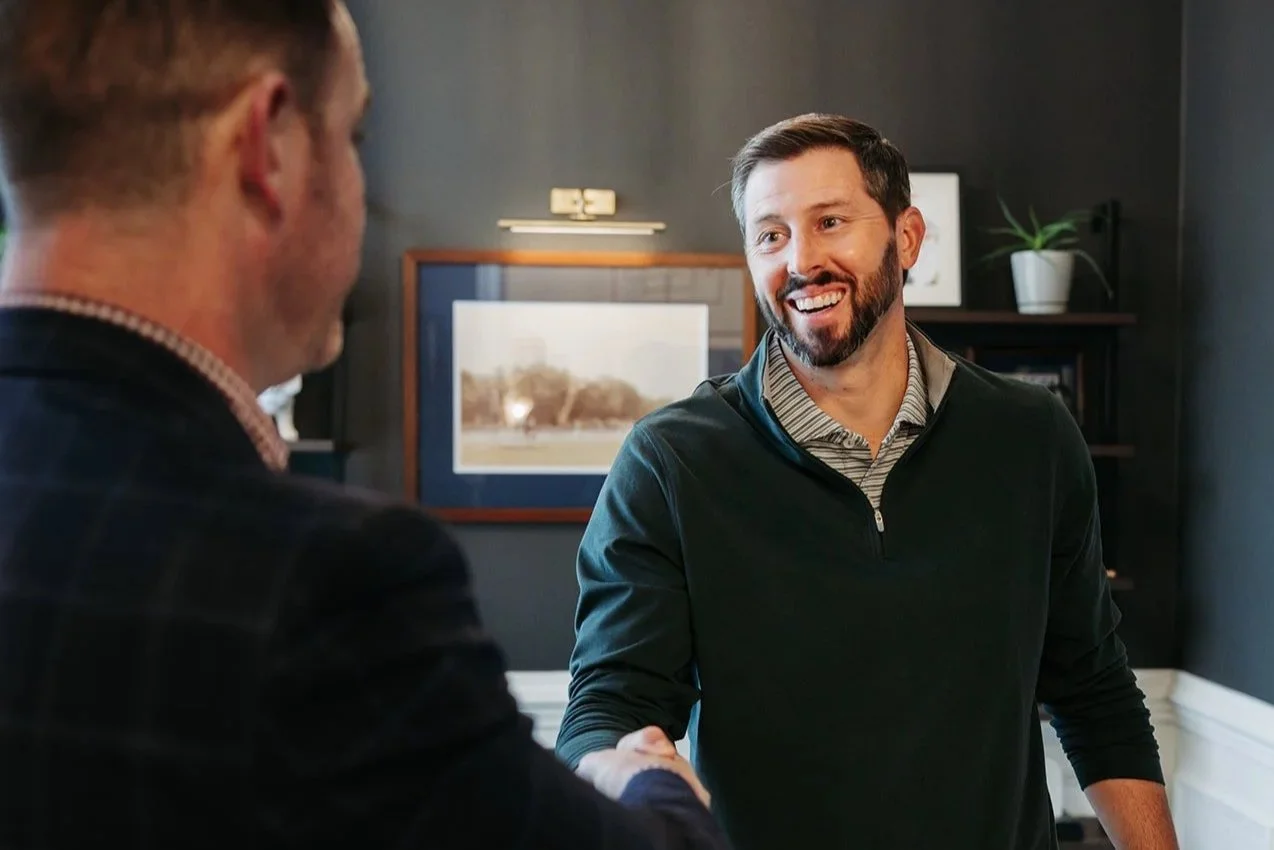 Two men shaking hands indoors, one smiling and facing the camera, the other with his back to the camera. There are framed pictures and a potted plant on a bookshelf behind them.