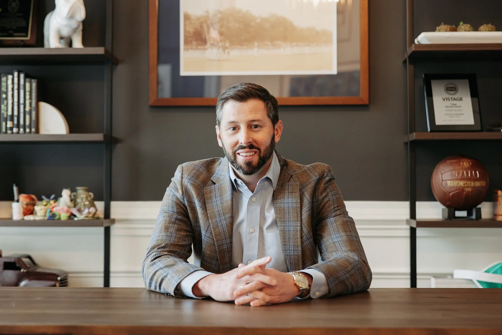 A man with dark hair and a beard sitting at a wooden desk with his hands clasped, wearing a checkered blazer and a white shirt, in an office with shelves and framed pictures in the background.