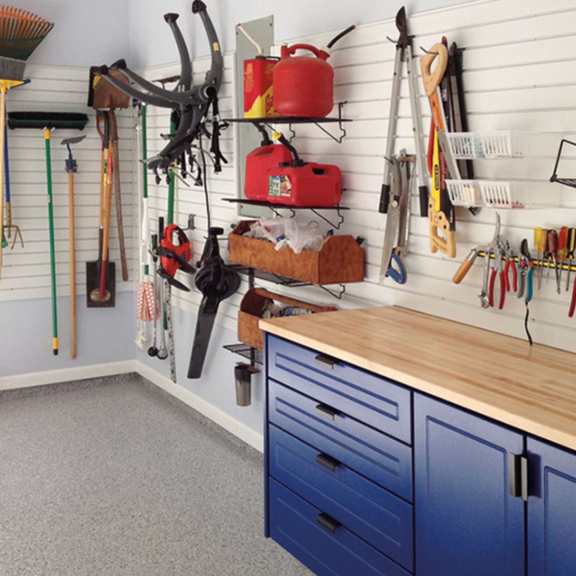 Garage workspace with a white slat wall system holding tools, equipment, and storage shelves beside blue cabinets with a wood countertop.