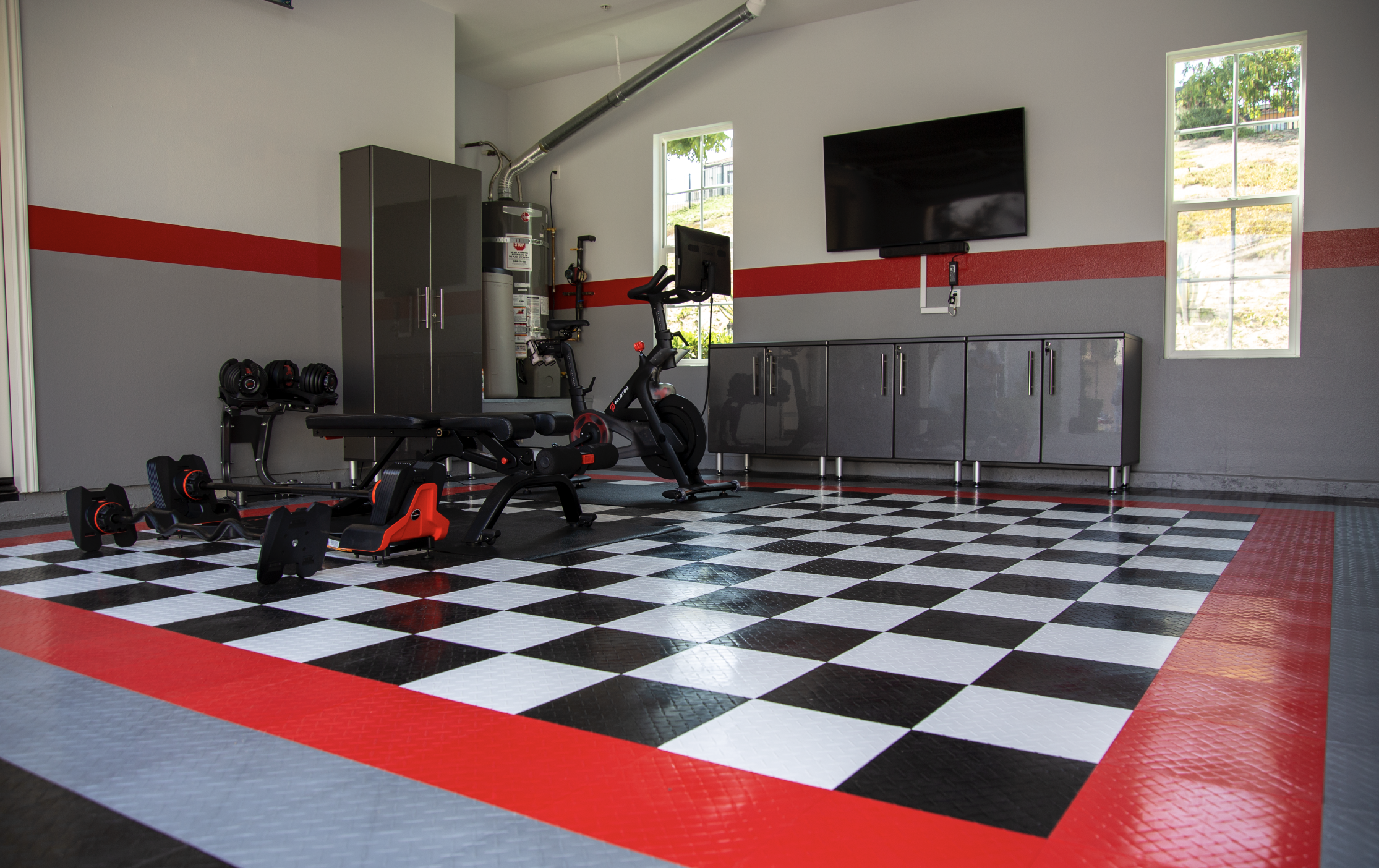 Garage gym with workout equipment including dumbbells, a weight bench, an exercise bike, and a punching bag, on a black and white checkered floor with red border, gray cabinets, a wall-mounted TV, and two small windows showing outdoor greenery.