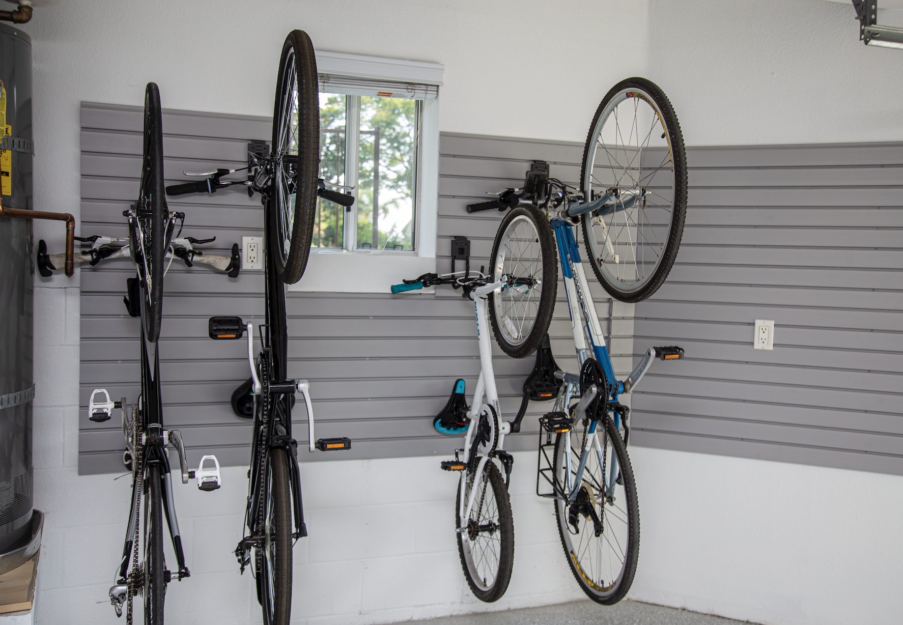 Garage with four bicycles mounted on a gray slatwall, with a window showing green trees outside.