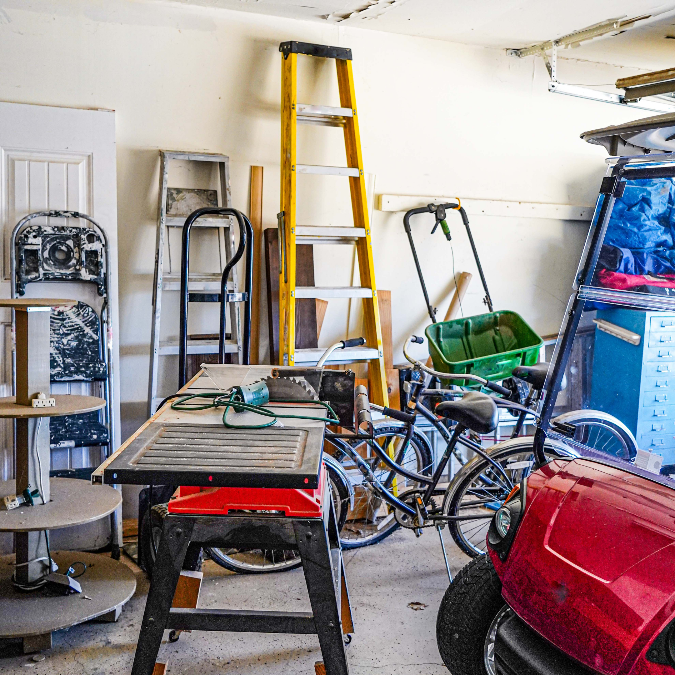 Cluttered garage with ladders, bicycles, a workbench, and scattered tools and equipment.