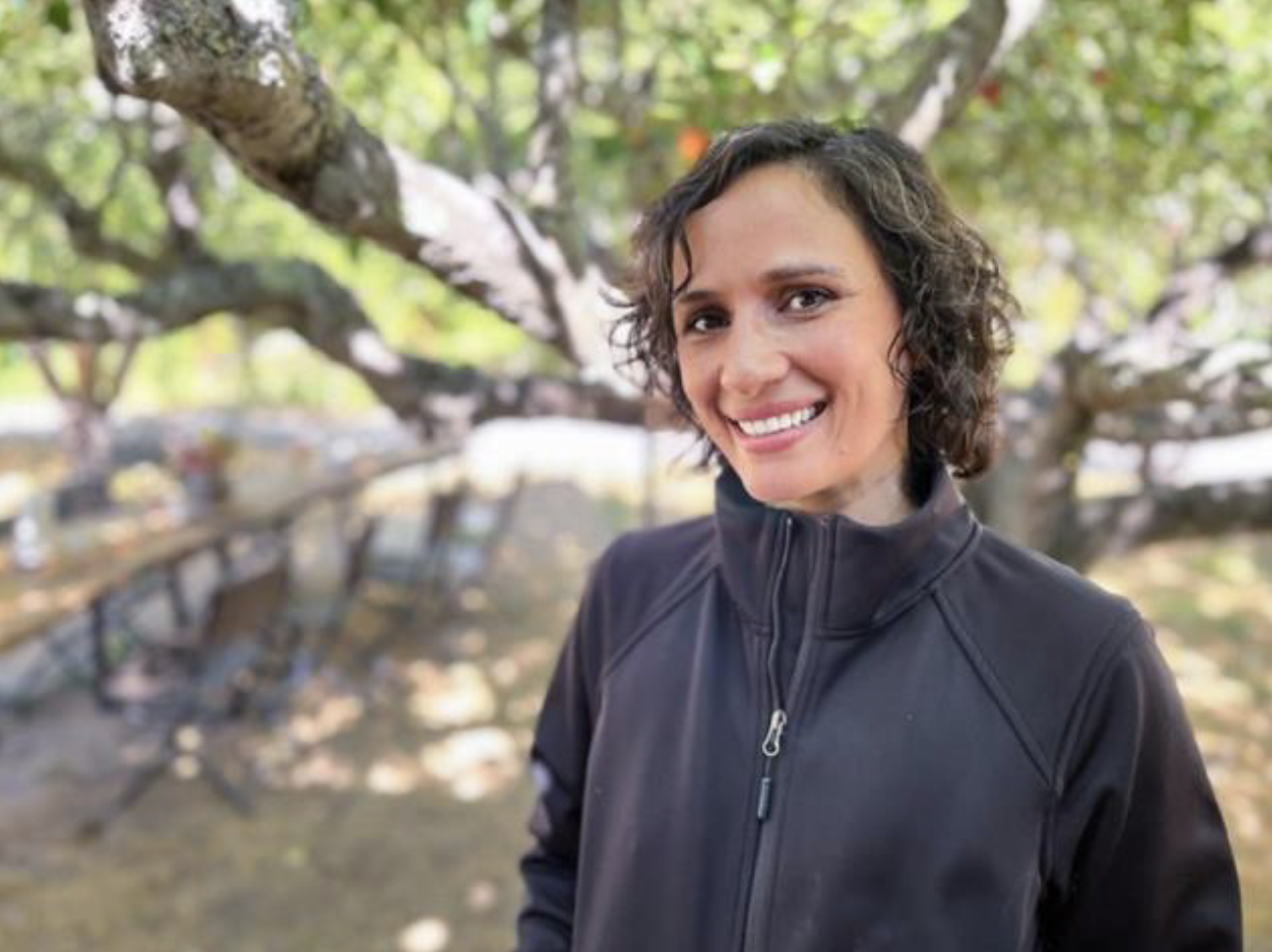 A woman with short curly hair wearing a black zip-up jacket, smiling outdoors with trees in the background.