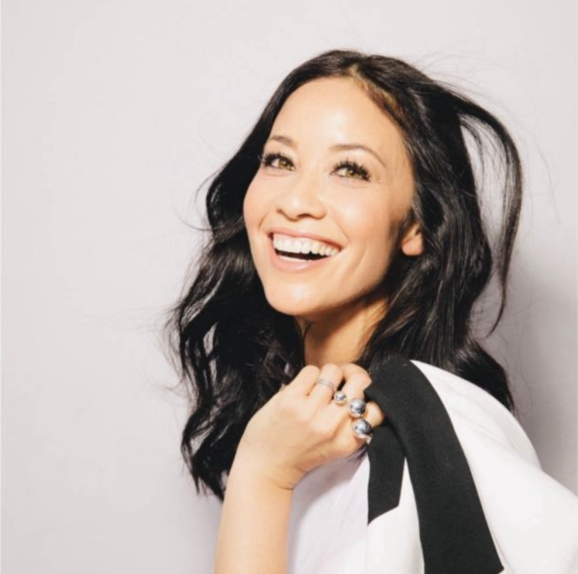 A woman with long dark hair smiling and looking to the side, wearing rings and a white top with a black shoulder detail.