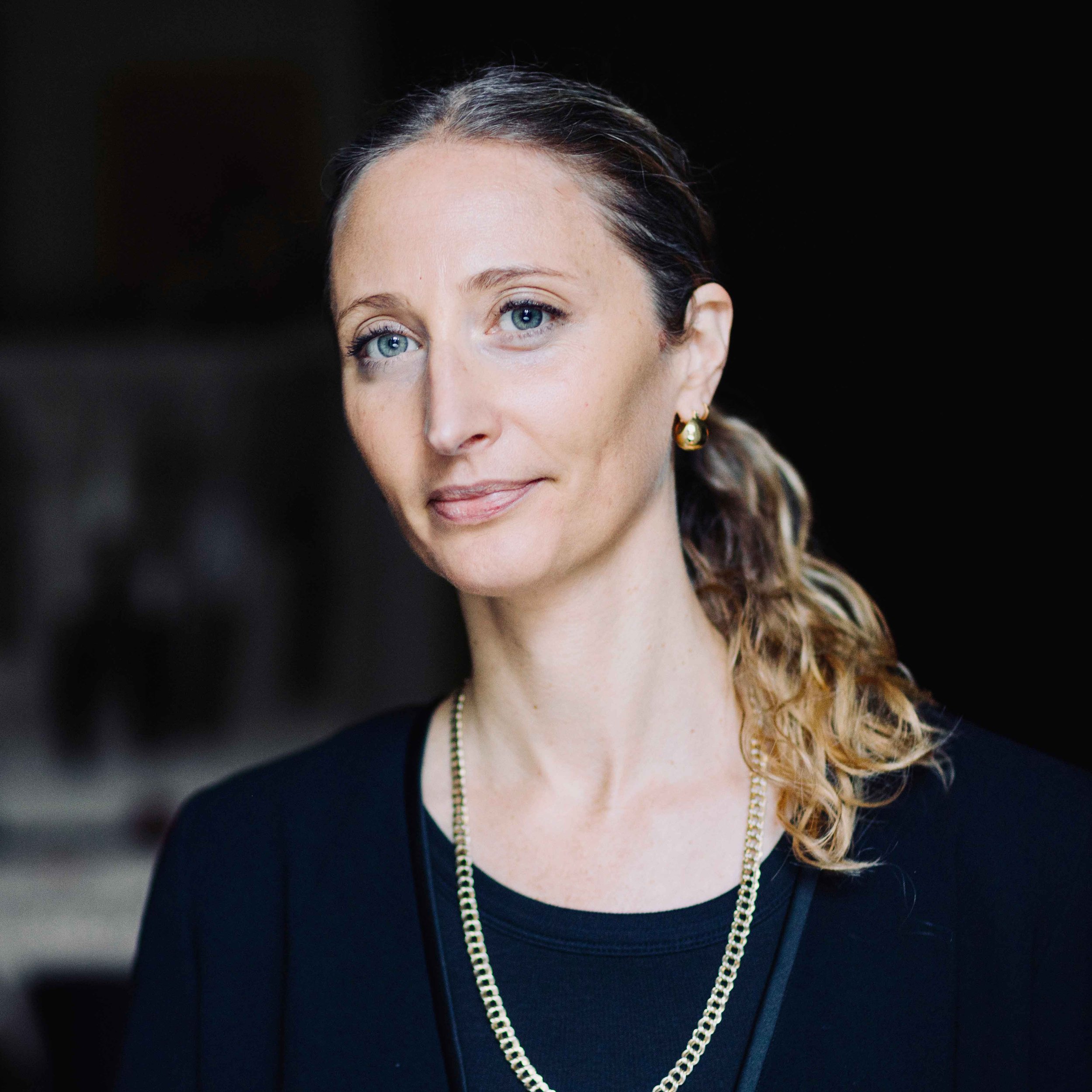 Portrait of a woman with blue eyes and light brown hair in a ponytail, wearing a black top, gold jewelry, and a neutral expression, against a dark background.