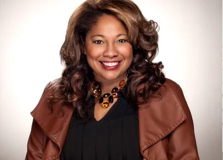Portrait of a woman with curly hair, smiling, wearing a black top and a brown jacket with a beaded necklace, against a white background.