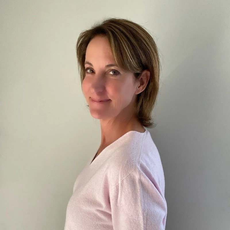 A woman with light brown hair smiling softly, wearing a light pink shirt, standing against a plain light-colored wall.
