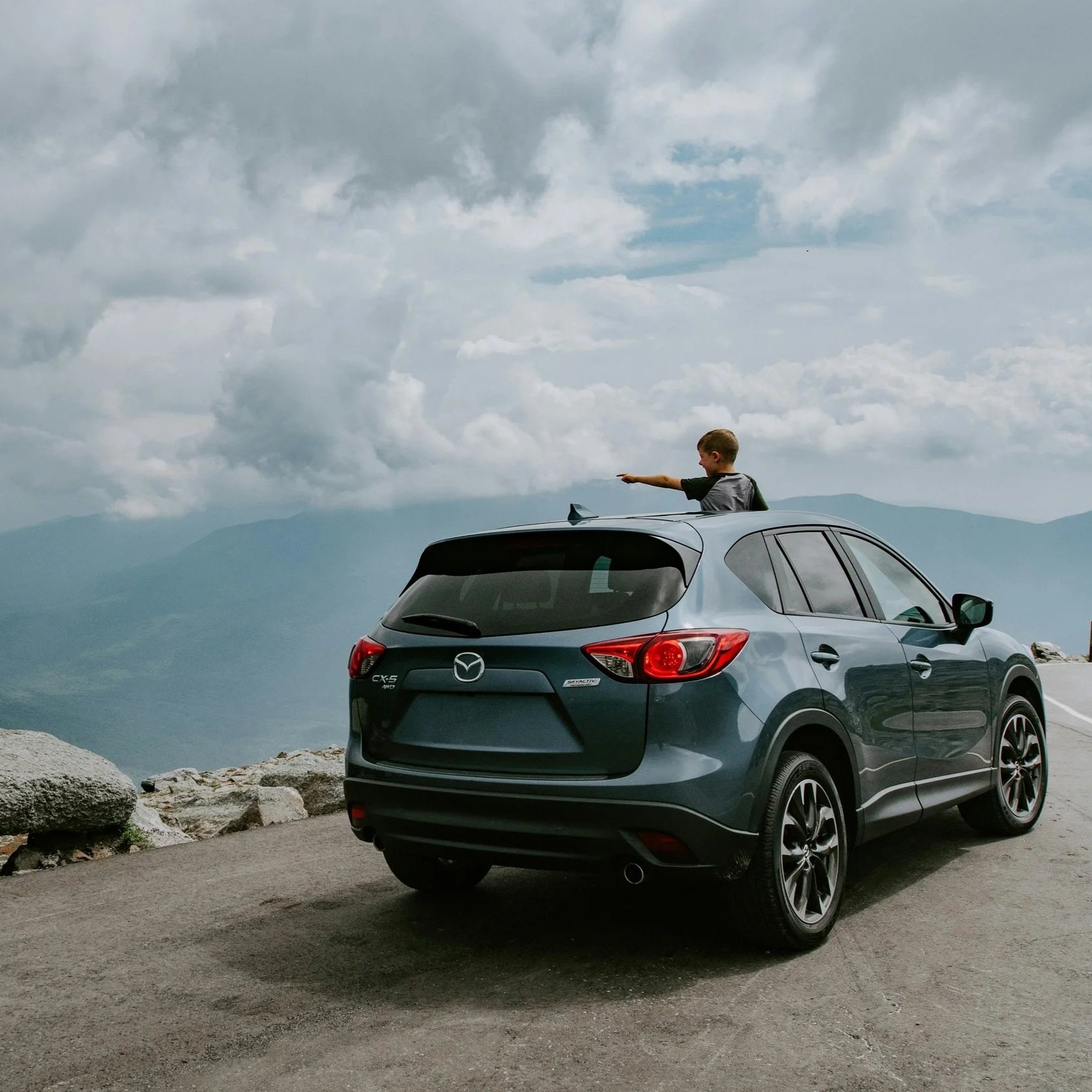 A child sticking out of the sunroof of a gray Mazda CX-5 parked on a mountain road, pointing towards the horizon with a mountainous landscape and cloudy sky in the background.