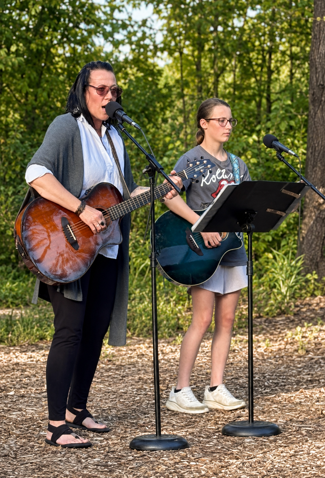 Two women, one older and one younger, performing music outdoors with guitars and microphones, surrounded by trees and greenery.