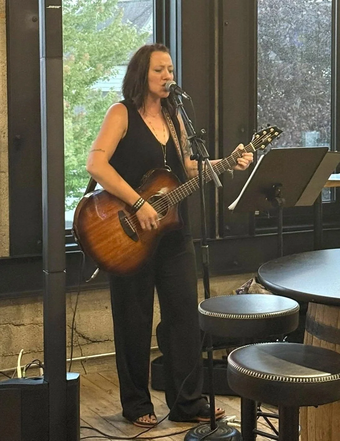 Amy Beach with dark hair wearing a black sleeveless top and black pants is singing and playing an acoustic guitar in a coffee shop or small venue. She stands in front of a window with trees outside.
