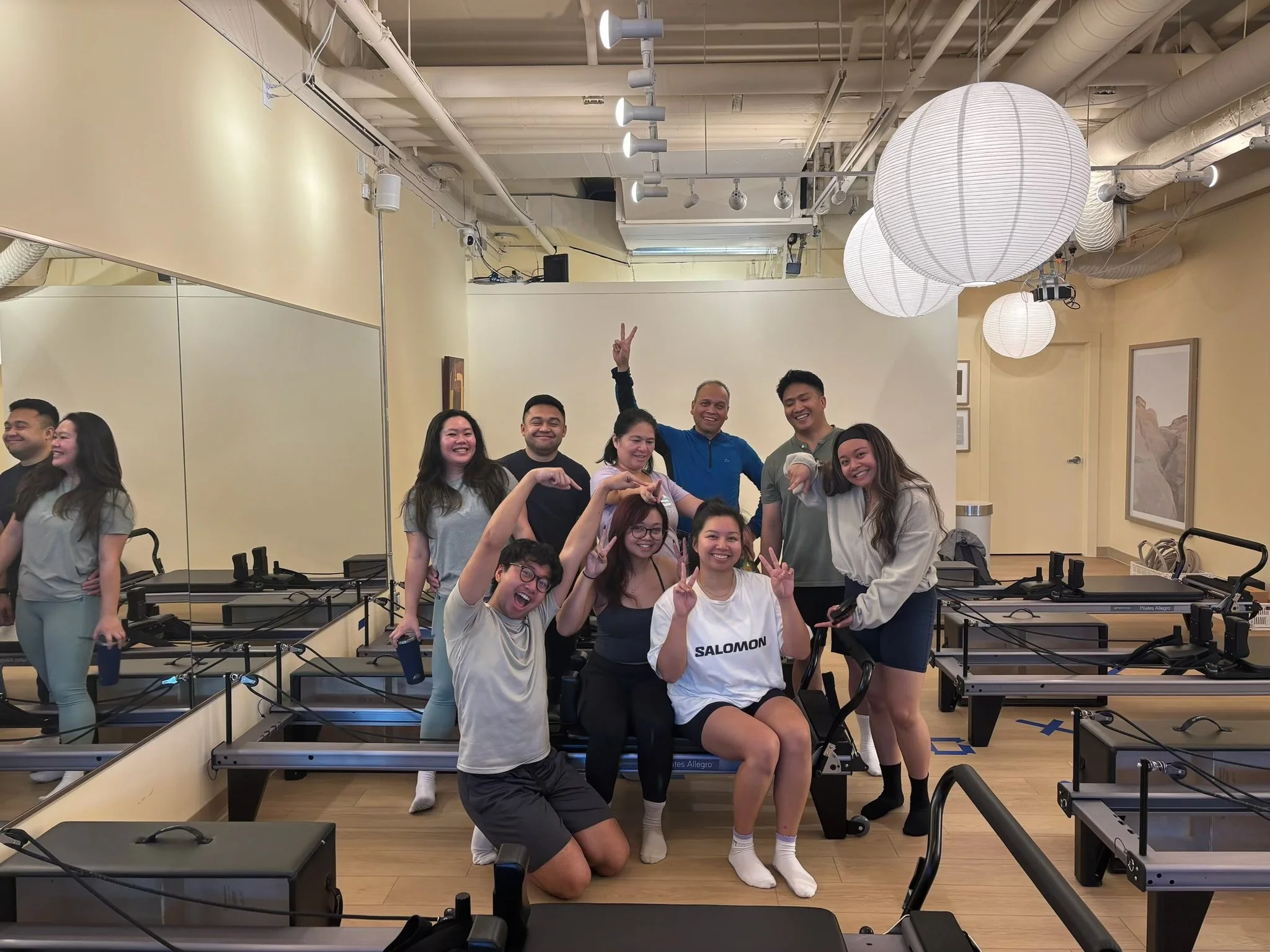 A group of people celebrating in a fitness class studio with pilates reformer machines, some making peace signs and smiling. The studio has a large mirror, soft lighting with large paper lanterns, and framed wall art.