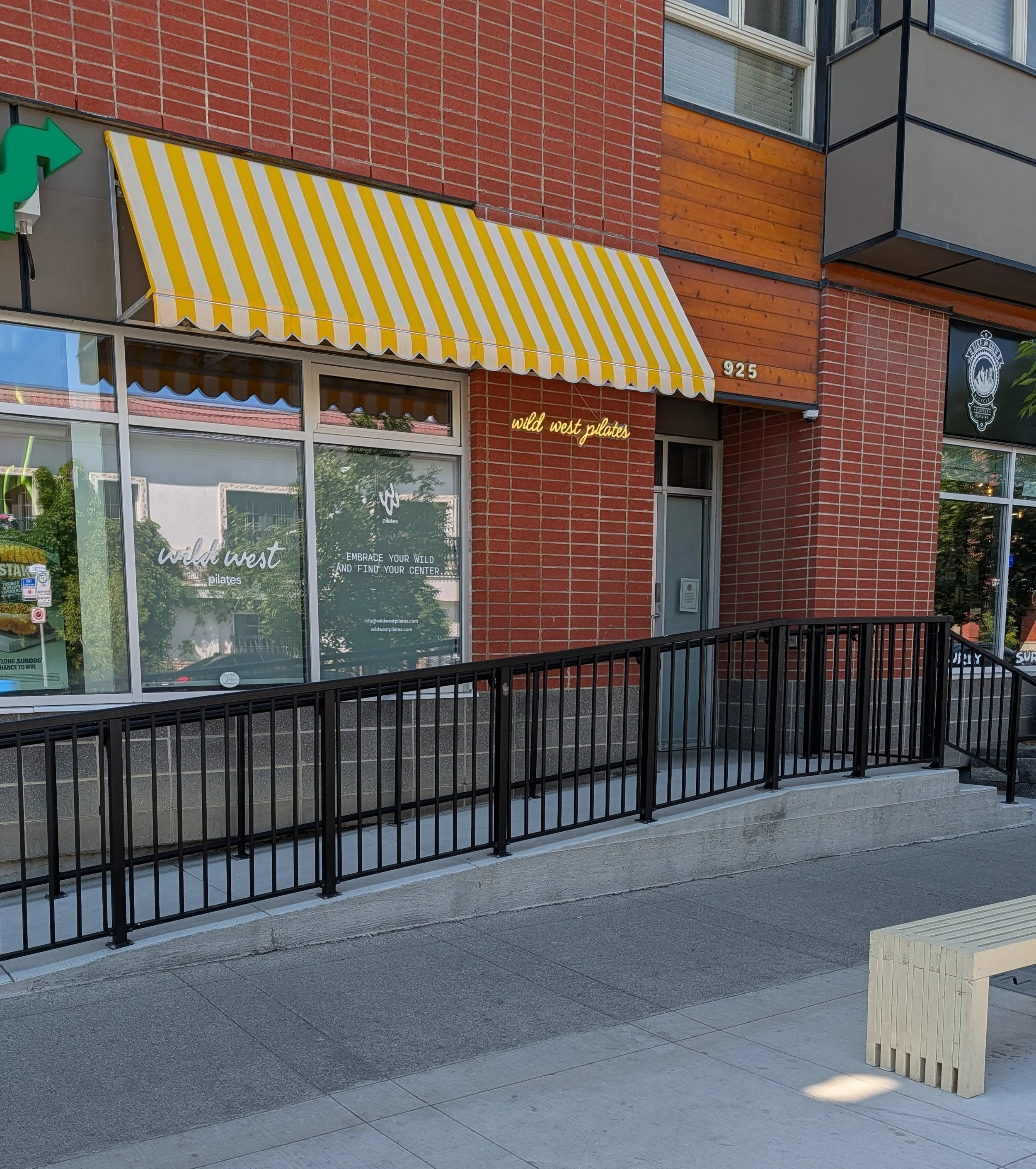 Exterior of a building with a yellow and white striped awning, a black railing, and a light wooden bench, with signage indicating business names 'wild west pilates' and a cannabis dispensary.